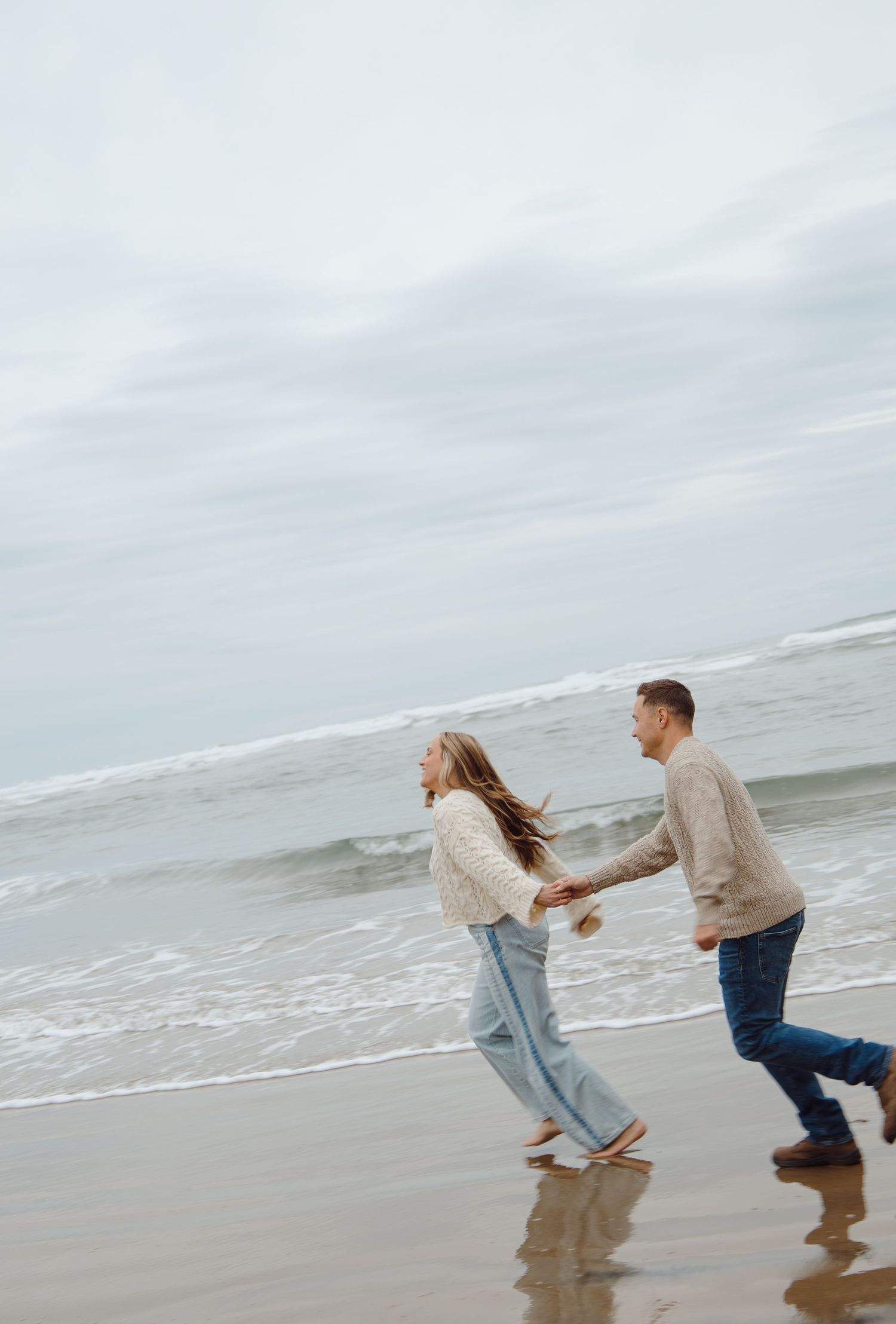 Couple running playfully along a wet sandy beach with ocean waves crashing in the background.