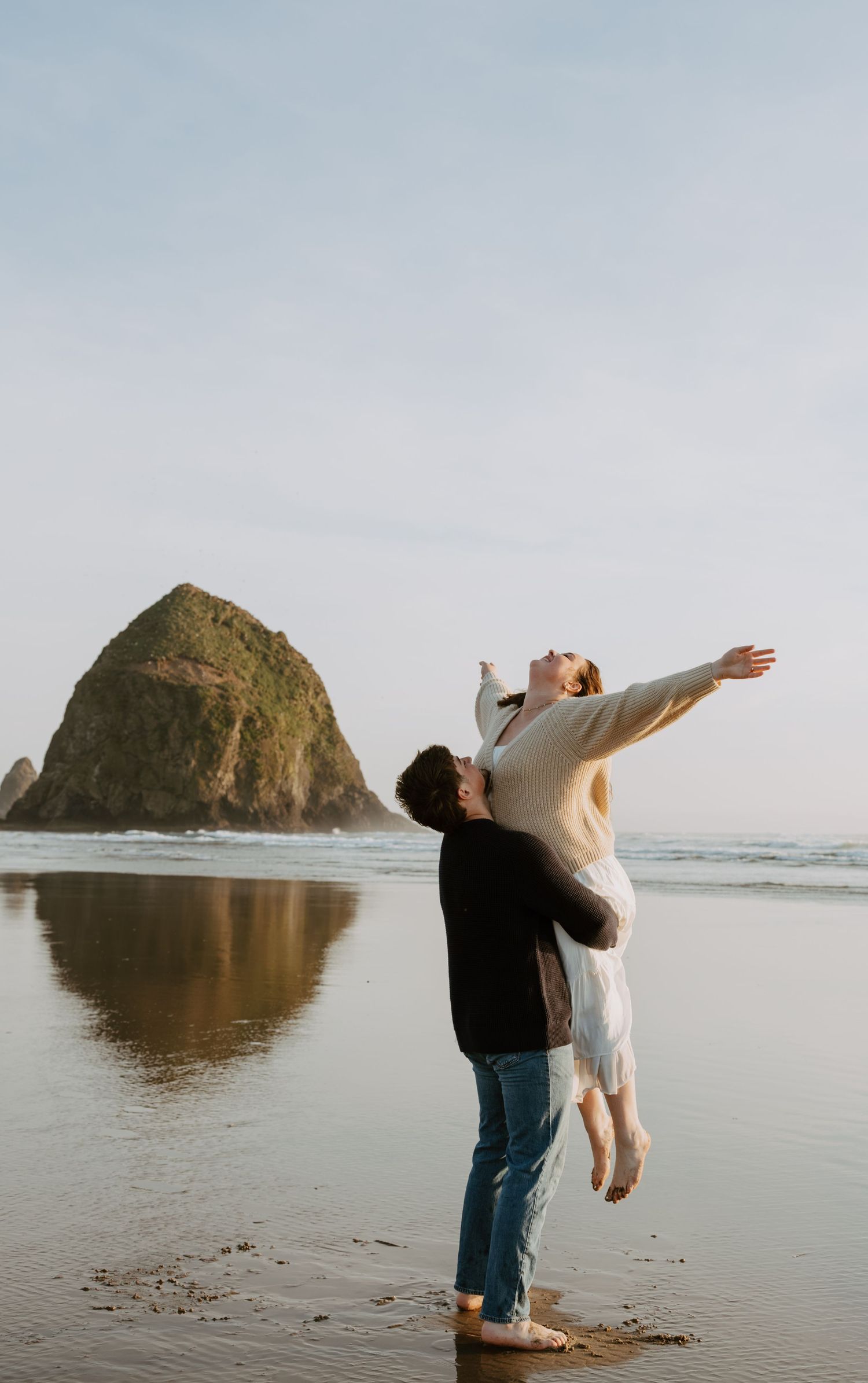 A couple shares a romantic moment on a beach near Haystack Rock, as a man lifts a woman with arms outstretched.