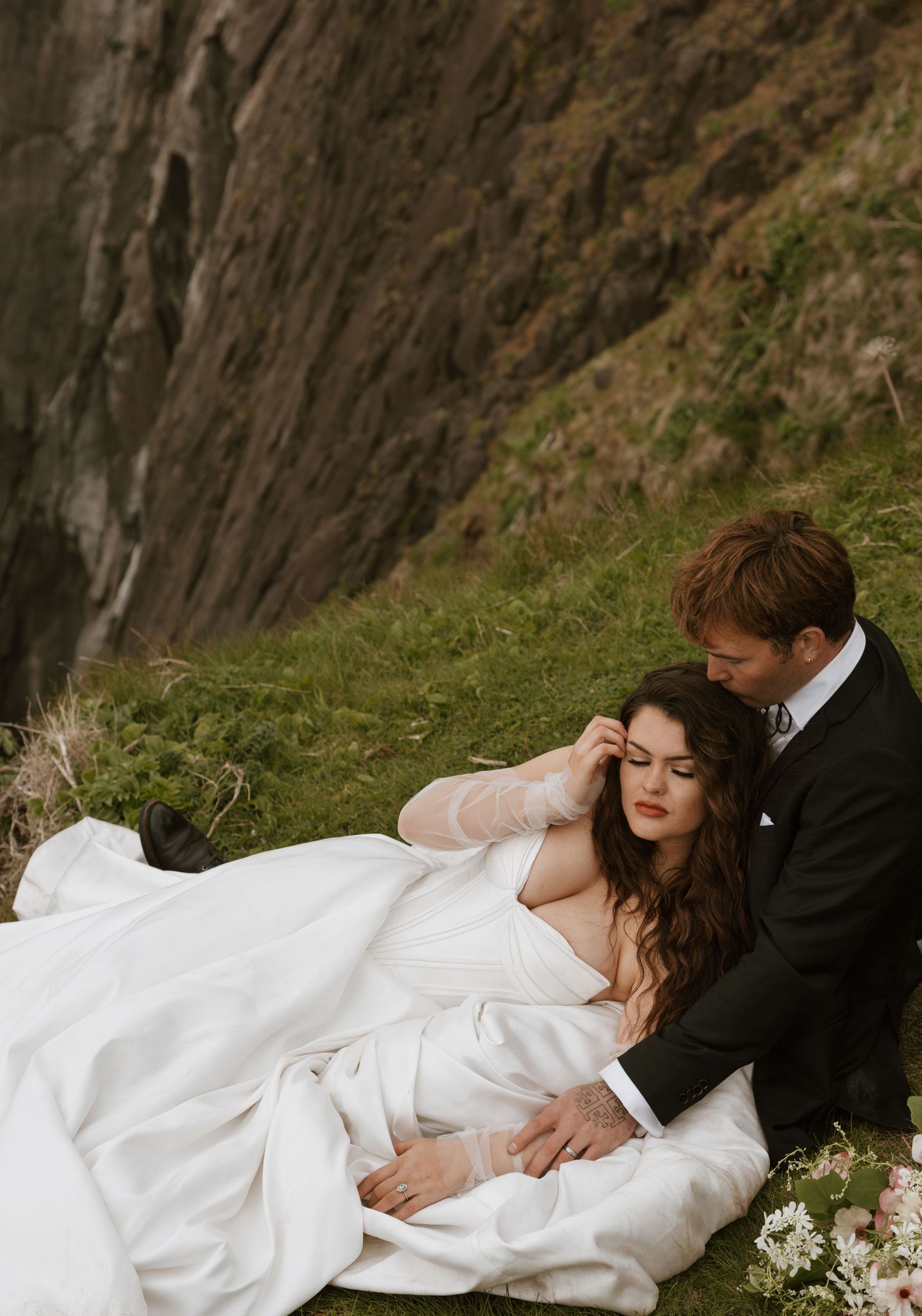 Bride in flowing white gown resting on groom in black suit on grassy cliffside with dramatic rock face behind.