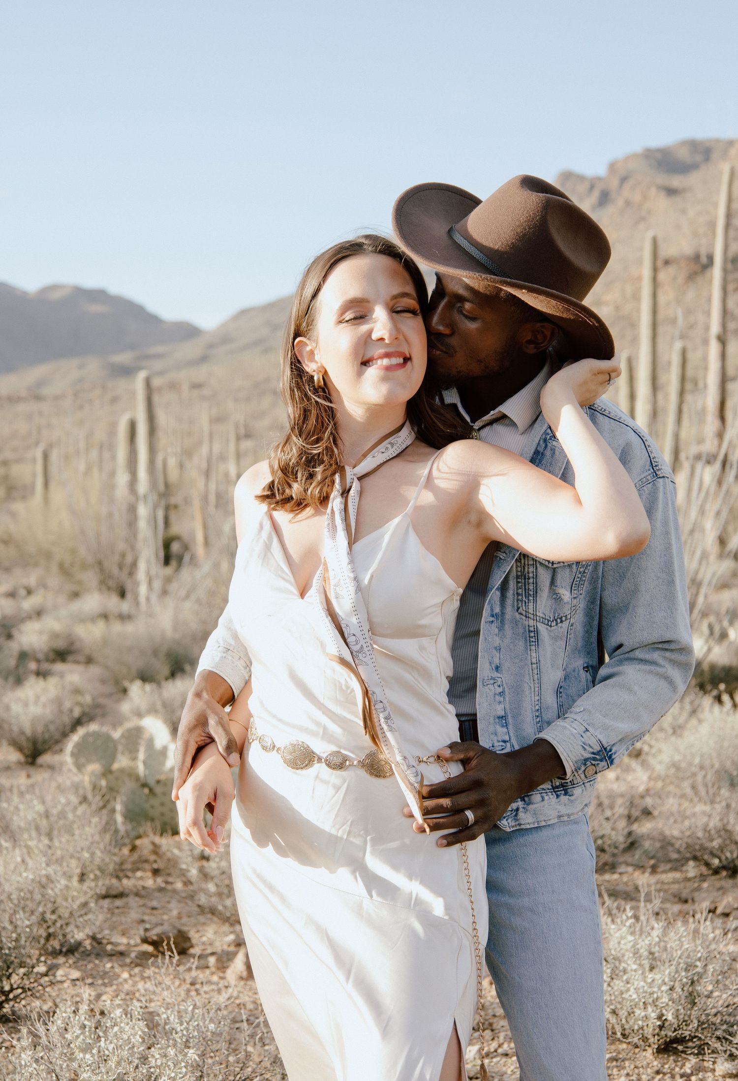 Man in cowboy hat kissing woman on cheek in desert setting, woman wearing white dress with shell belt accessory.