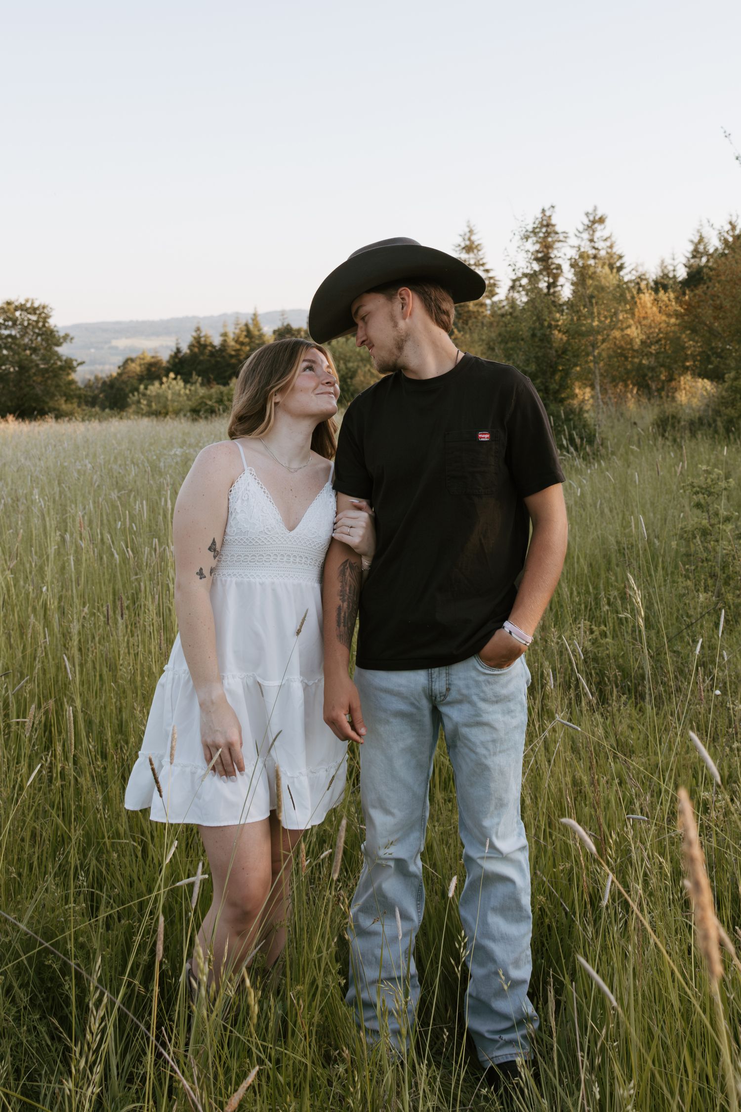 Young couple gazing at each other in a lush green meadow, woman in white dress, man in black tee and cowboy hat.