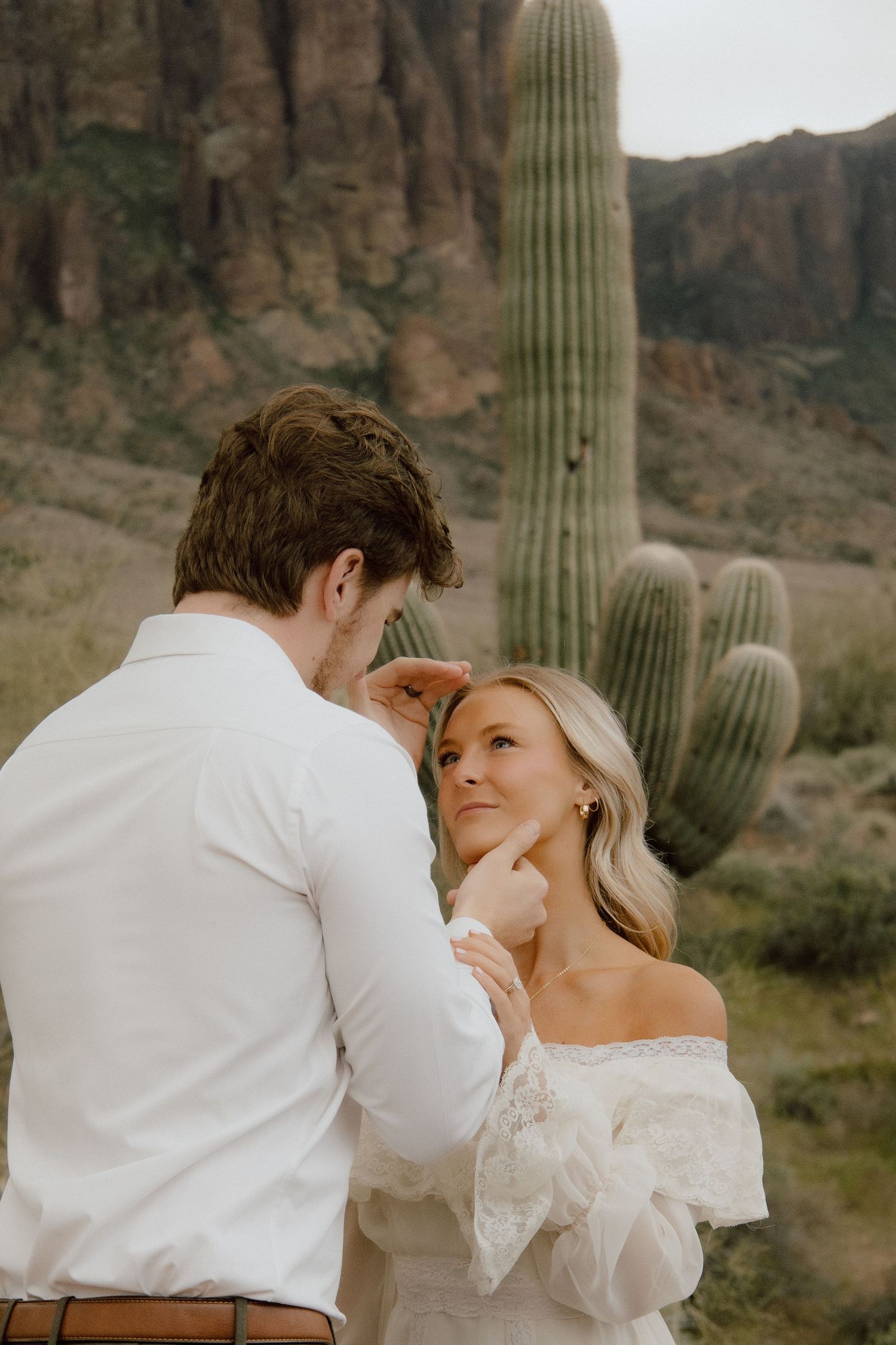 Couple shares tender moment in desert, woman in off-shoulder lace dress gazes up at man by cactus.