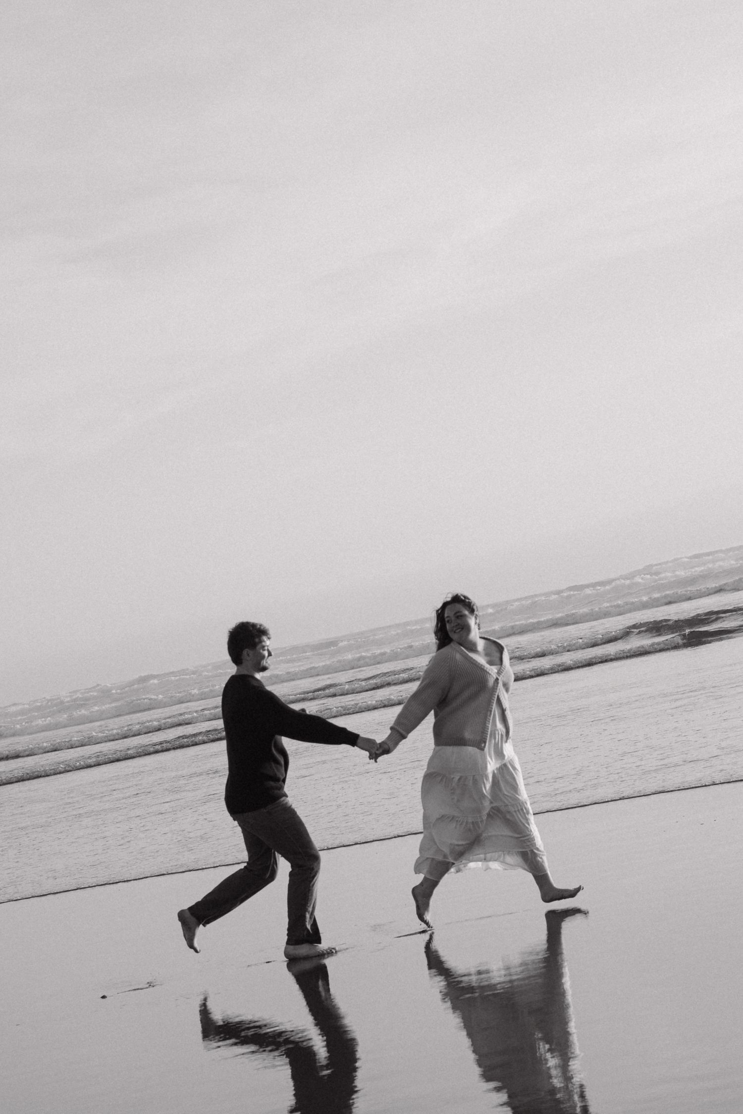 Black and white photo of a couple holding hands and running barefoot on a reflective wet beach shoreline.