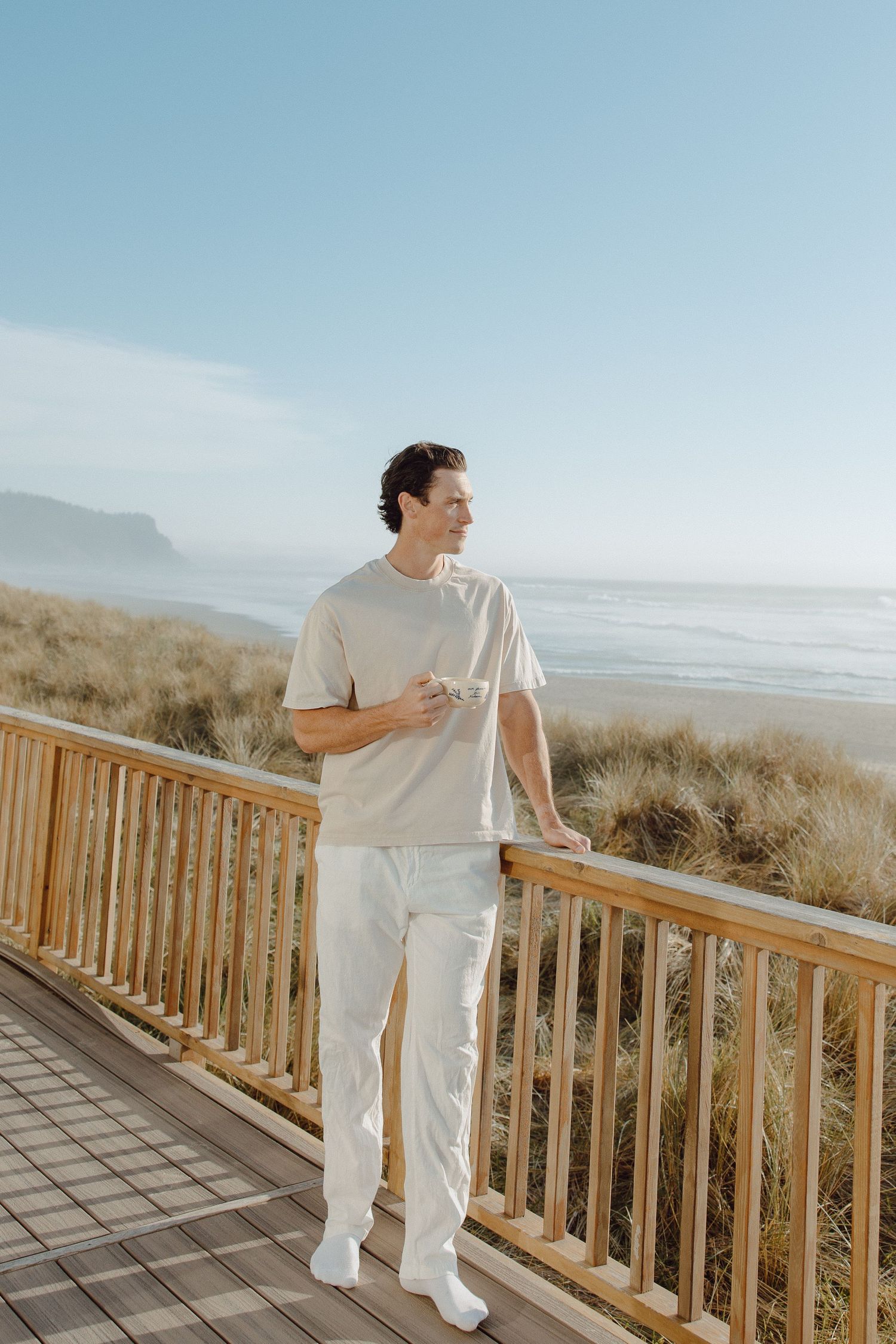 Man in beige t-shirt and white pants standing on a wooden deck overlooking a scenic beach on a clear day.