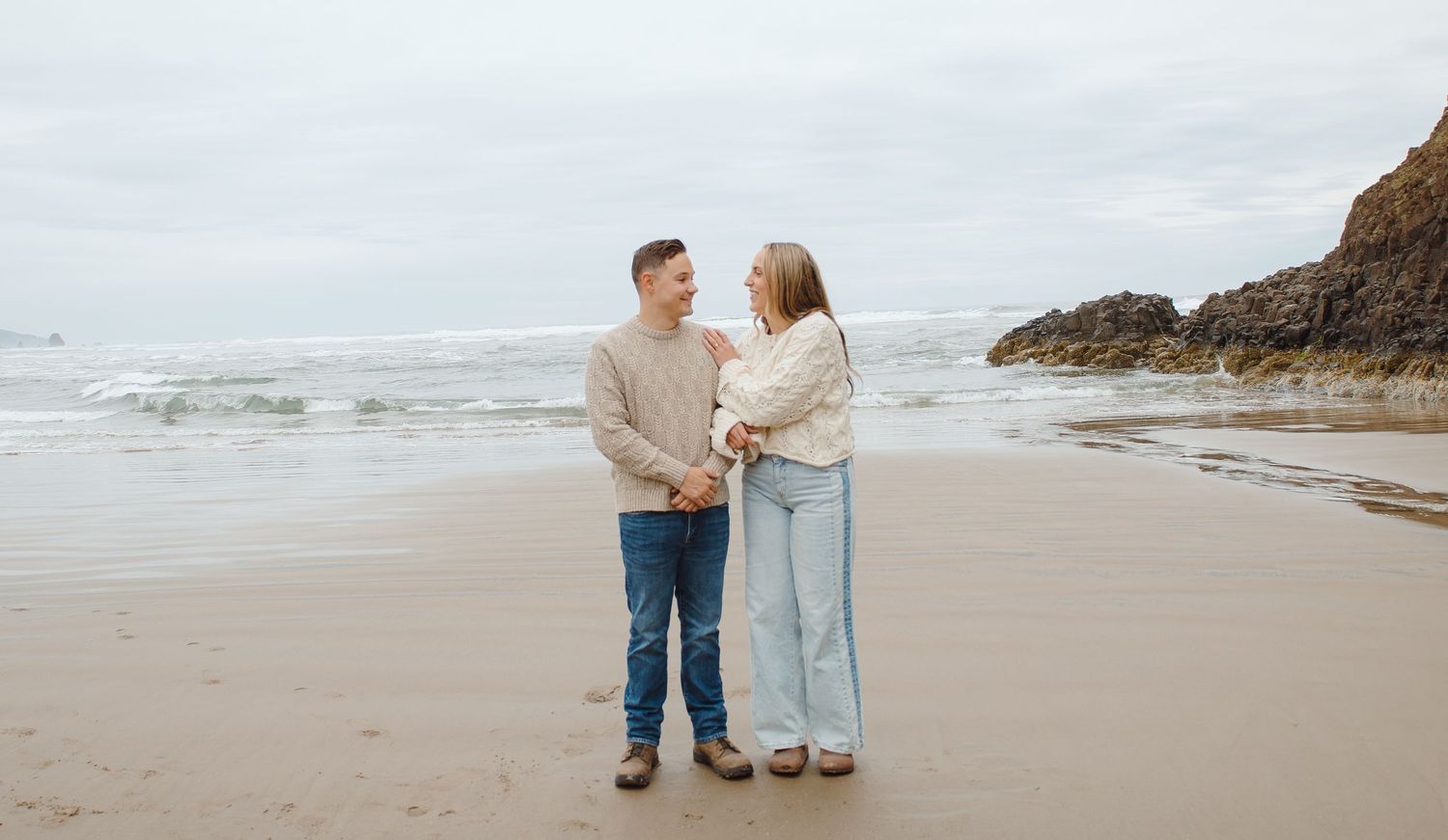 Happy couple standing together on a sandy beach holding hands with ocean waves and rocks behind them.