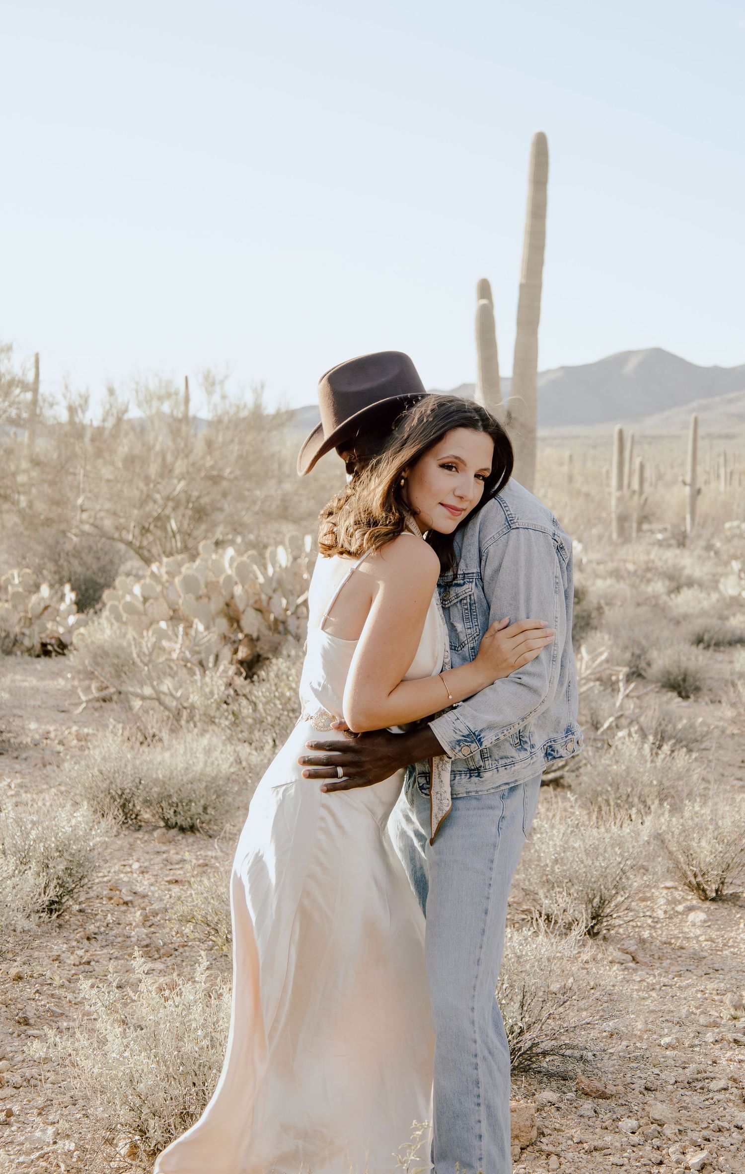 Woman in white dress embracing man in cowboy hat from behind among saguaro cacti in Arizona desert landscape.