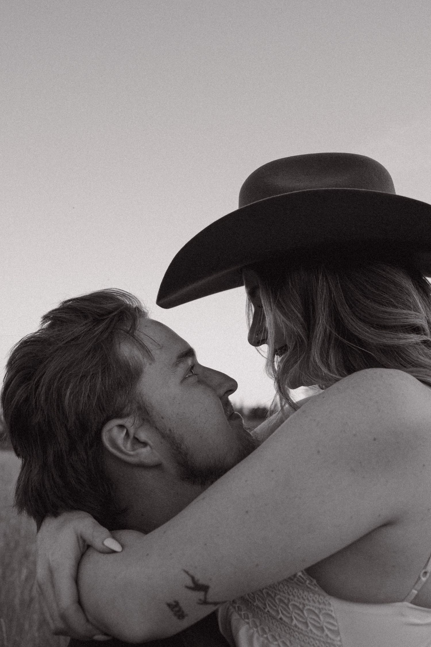 Black and white photo of a couple embracing outdoors, woman wearing a cowboy hat leaning in for a kiss.