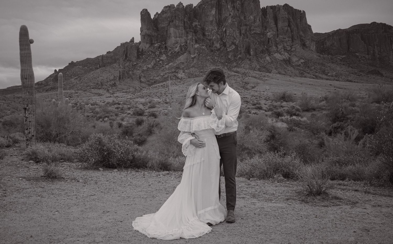 Black and white photo of bride and groom embracing in desert landscape with rocky mountain backdrop.