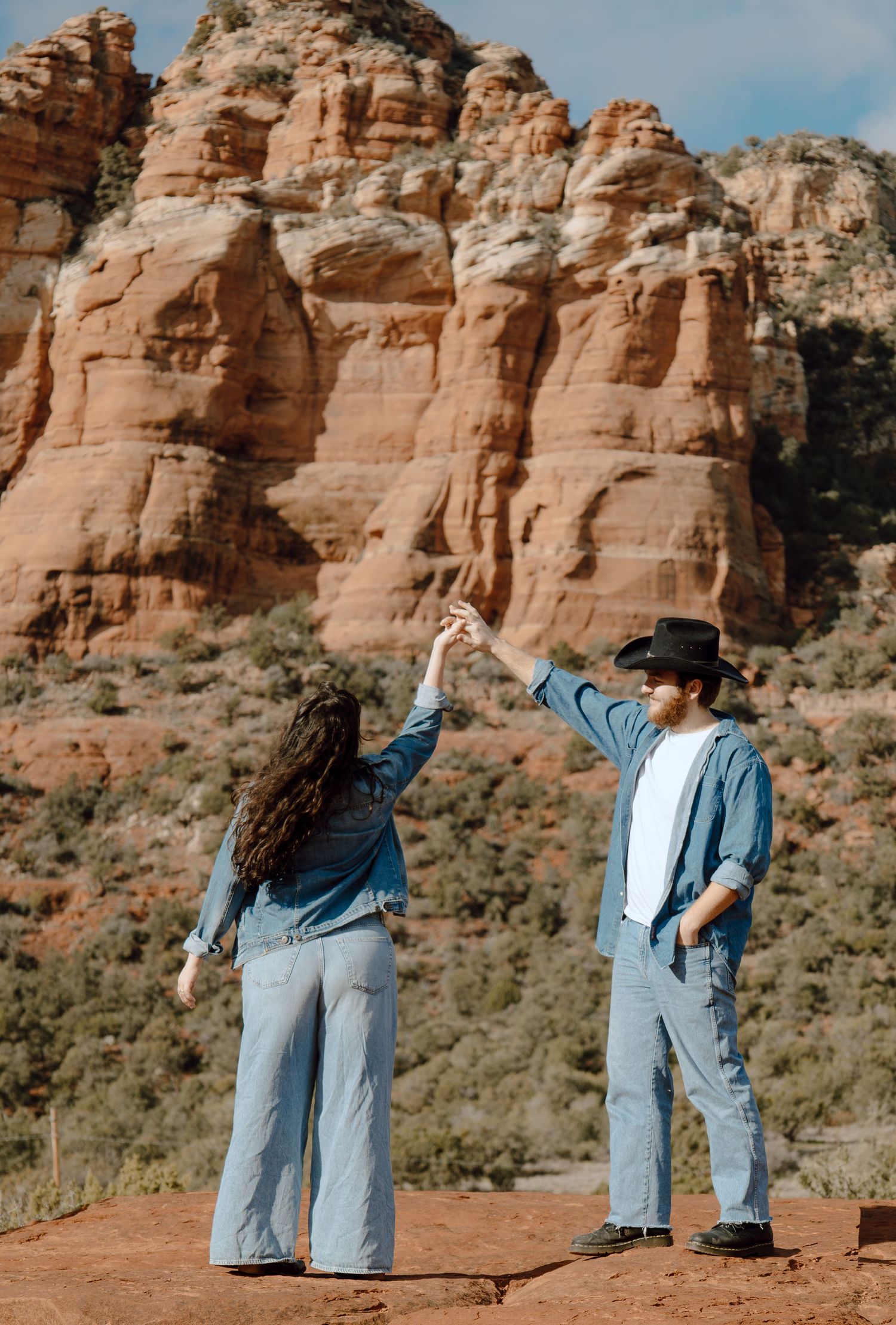 Couple holding hands up in matching denim outfits against red rock formations in Sedona, Arizona.