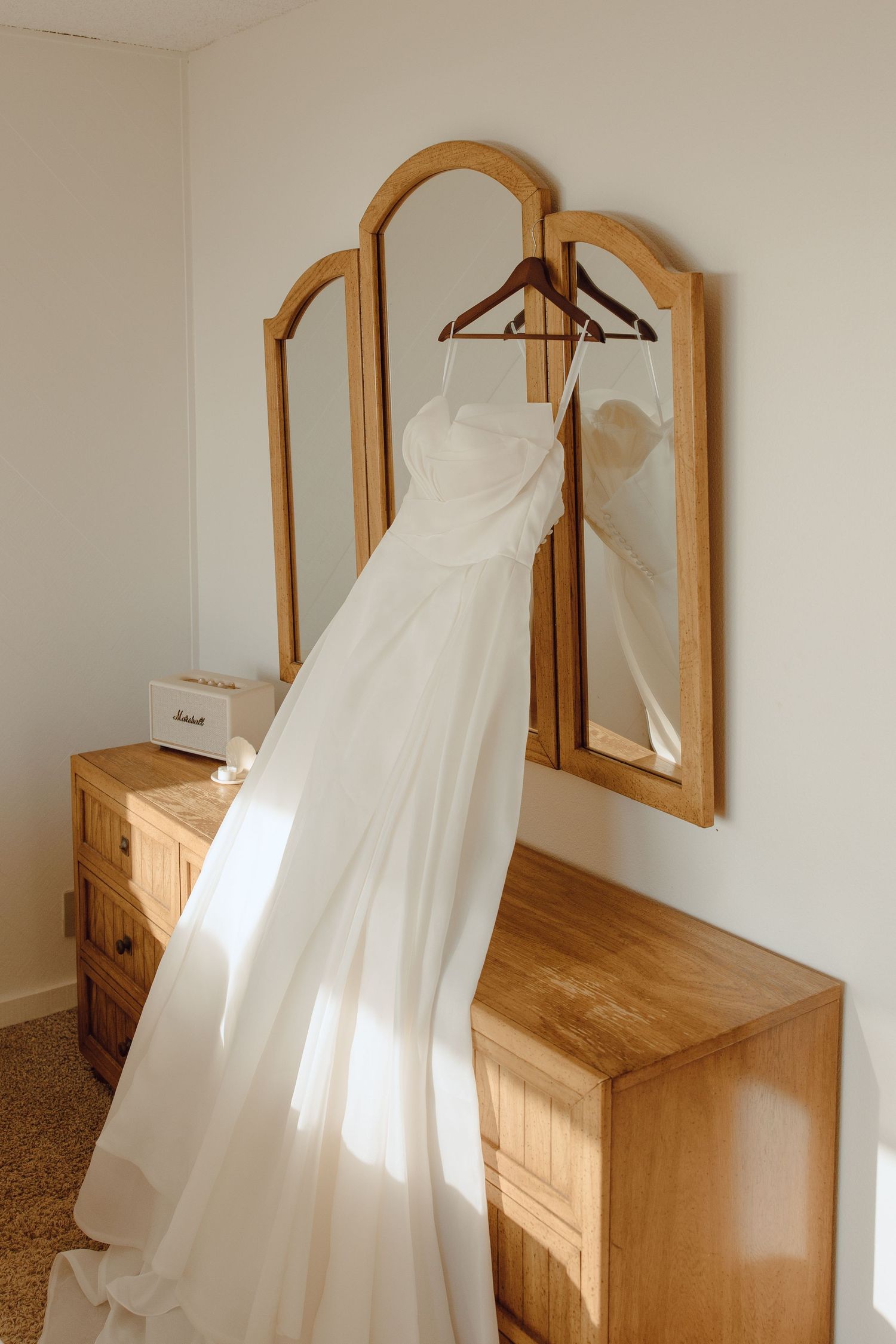 White wedding dress hanging on a wooden mirror dresser in a sunlit room.