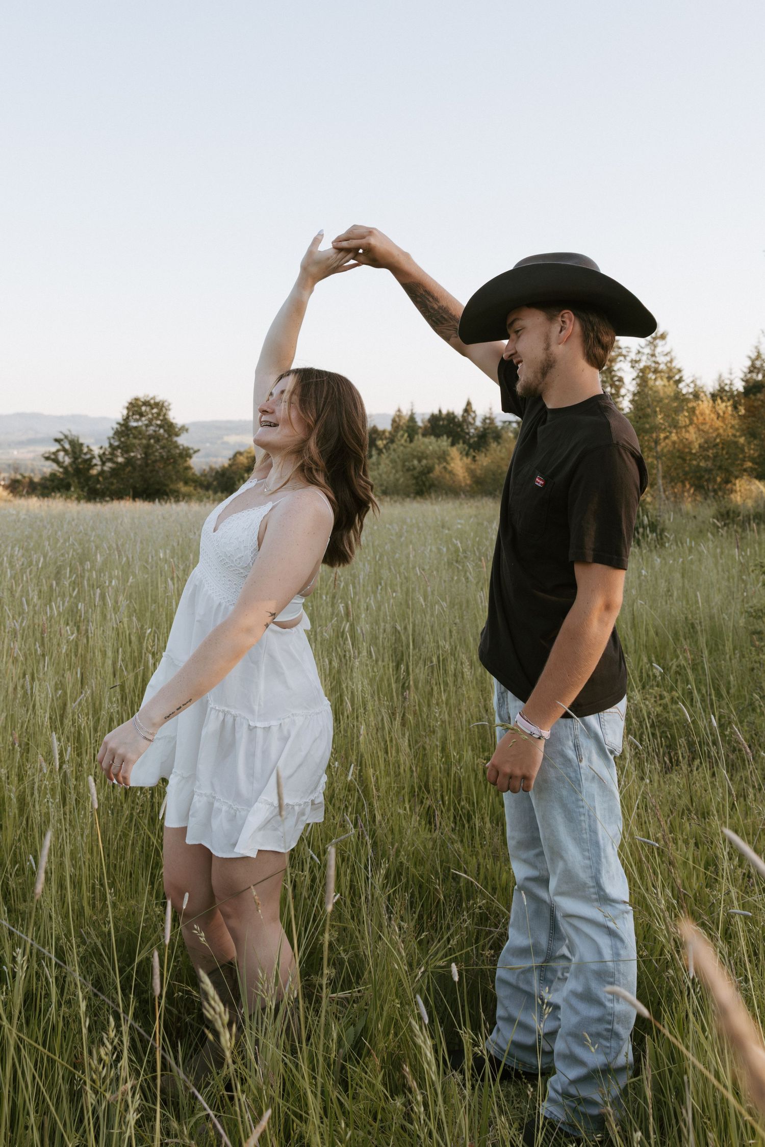 Couple dancing in a green field, woman in white dress twirling, man in black tee and cowboy hat, golden hour.