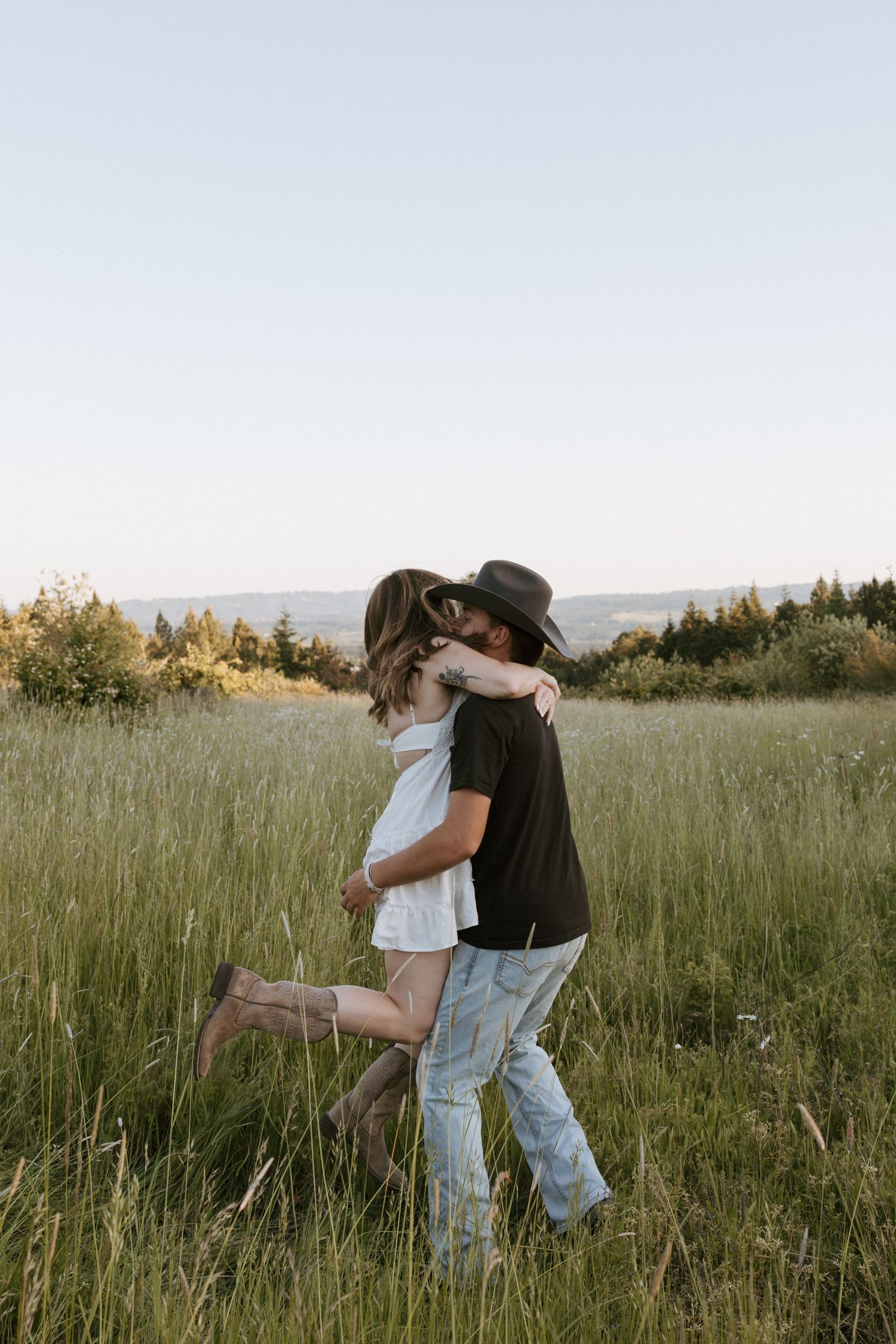 Couple embracing and kissing in a lush green meadow, man wearing cowboy hat and jeans, woman in white dress.