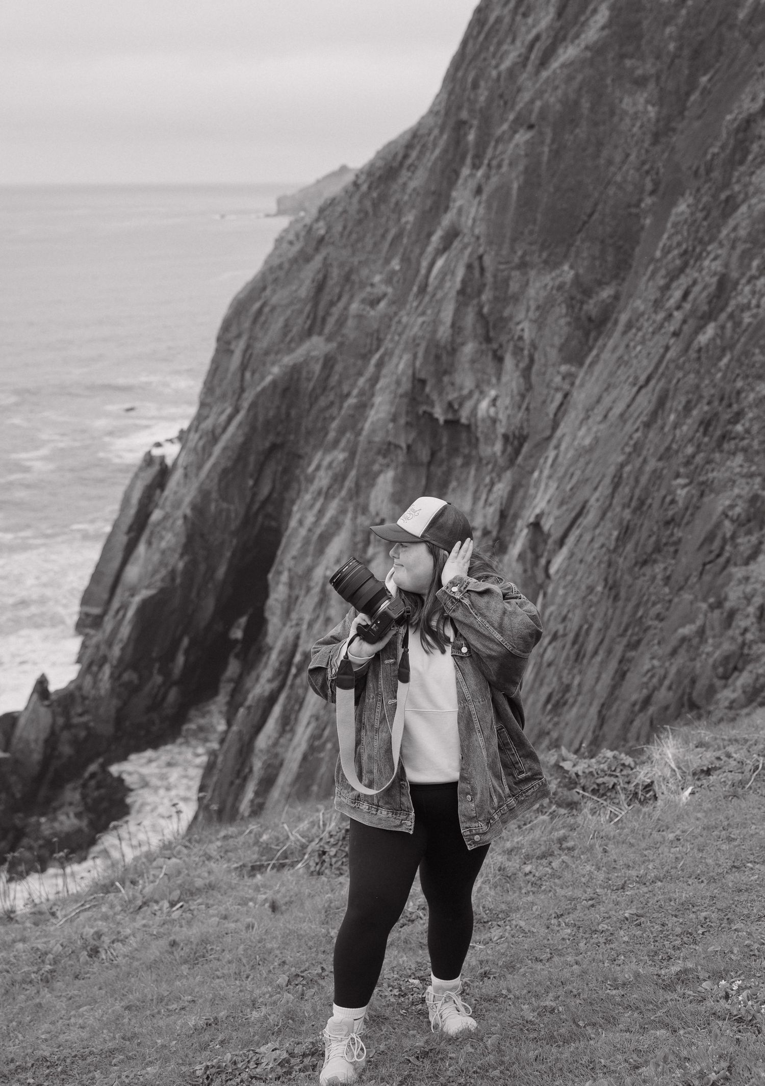 Black and white photo of a photographer with camera raised, standing on coastal cliffs with ocean views.