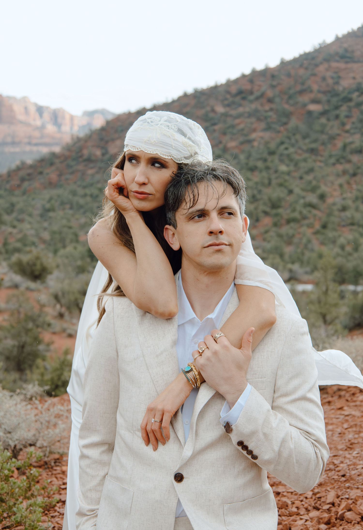 Bride in white headscarf leans on groom's shoulder against Sedona red rock background.