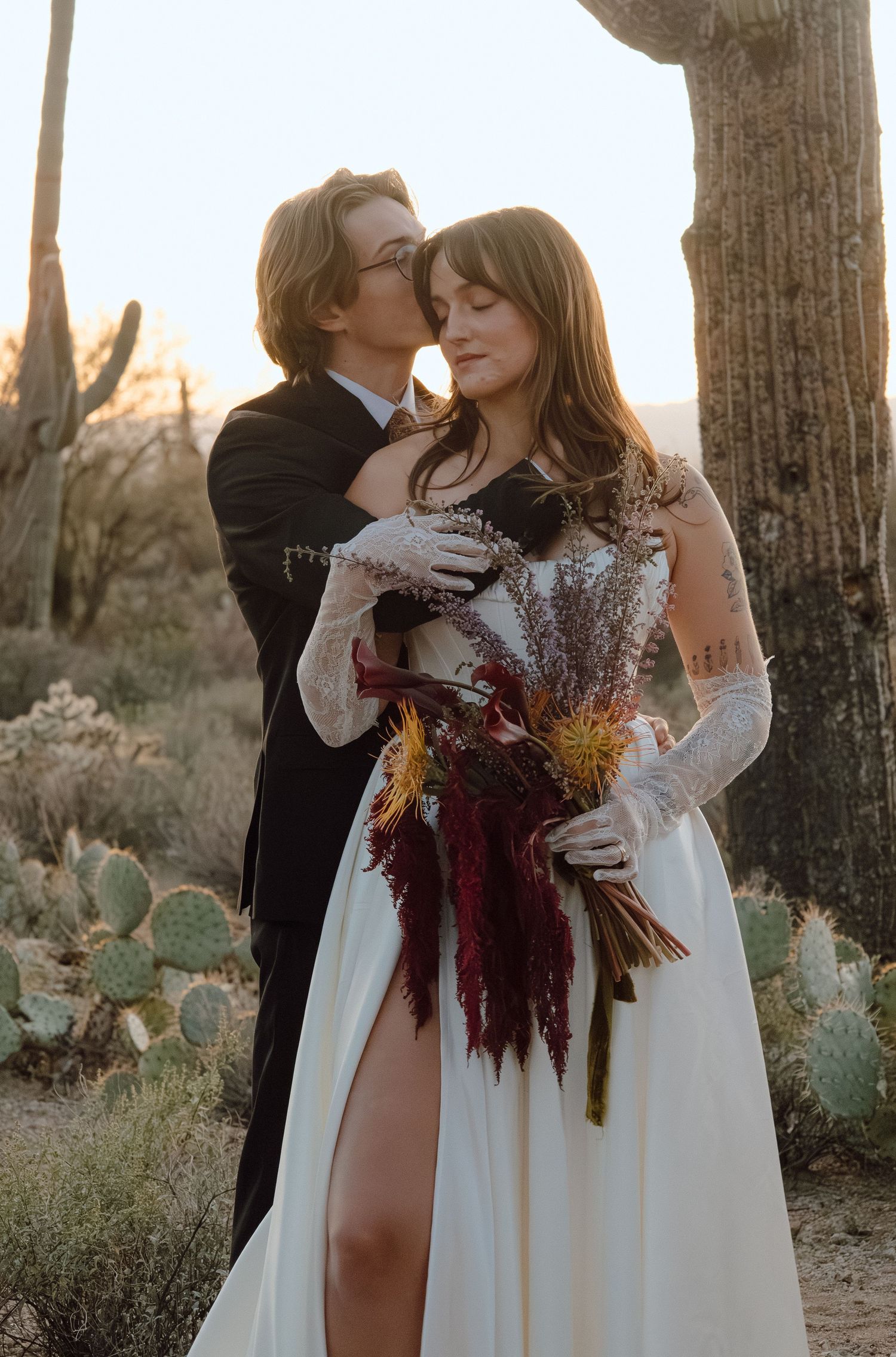 Groom kisses tattooed bride on cheek as she holds a boho dried floral bouquet among desert cacti.