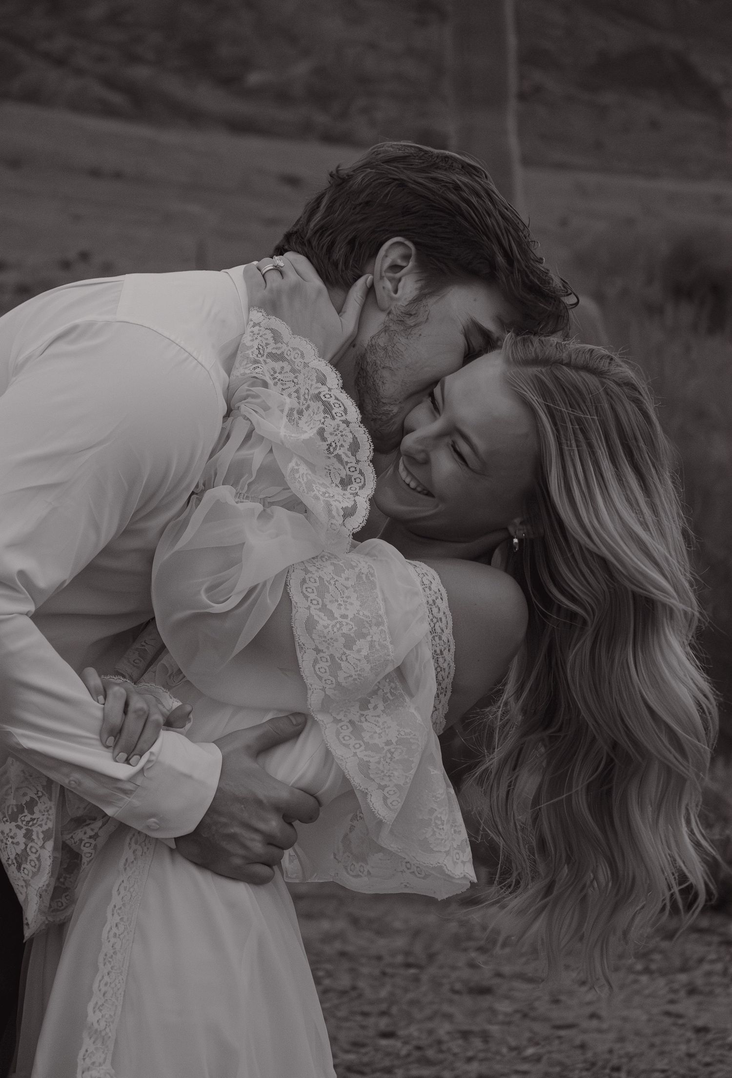 Black and white photo of man kissing woman on cheek as she laughs, embracing in desert landscape.