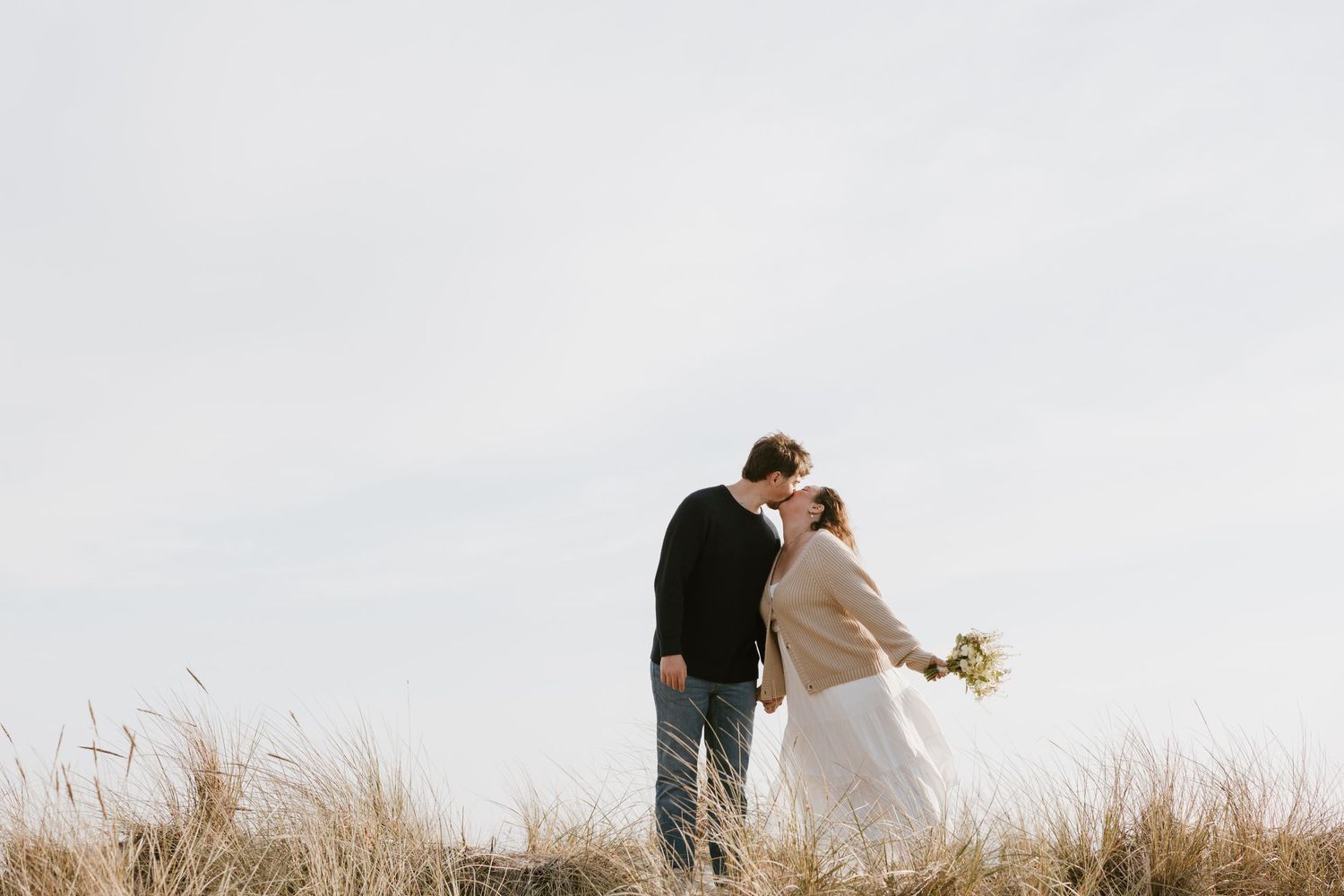 A couple shares a romantic kiss in a golden grass field, bride holding a bouquet, against a soft sky backdrop.