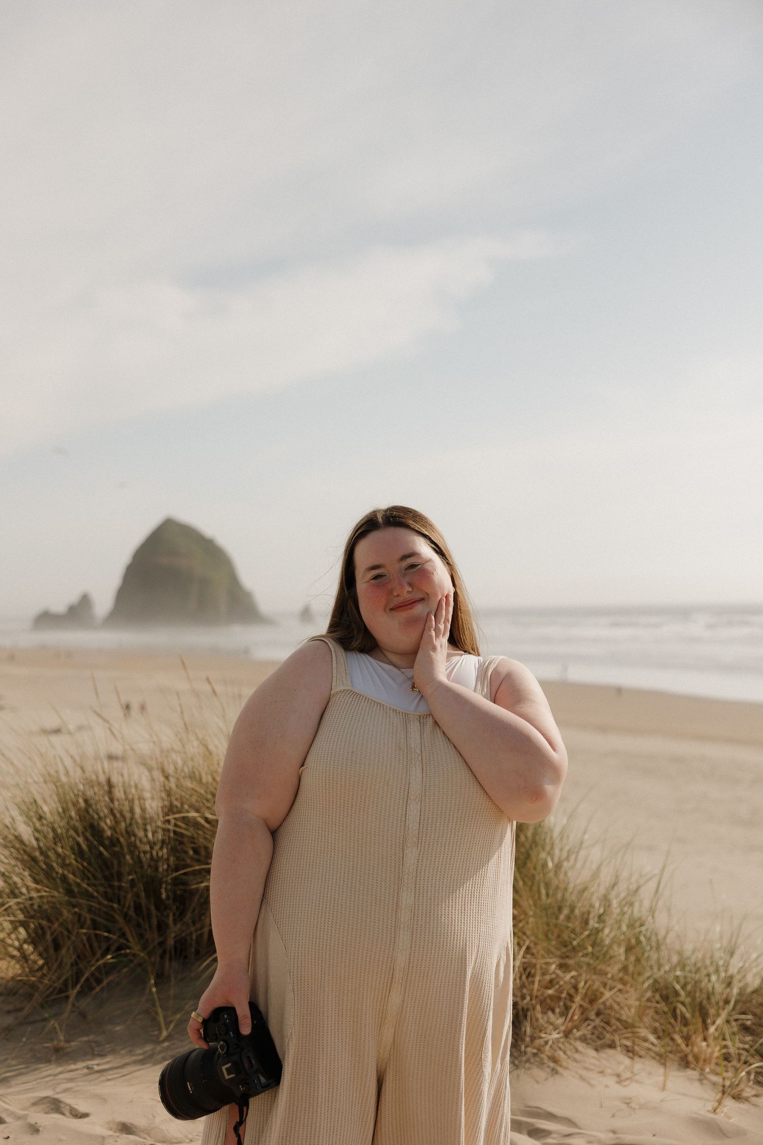 Woman in beige jumpsuit holding camera on sandy beach with Haystack Rock in background on cloudy day.