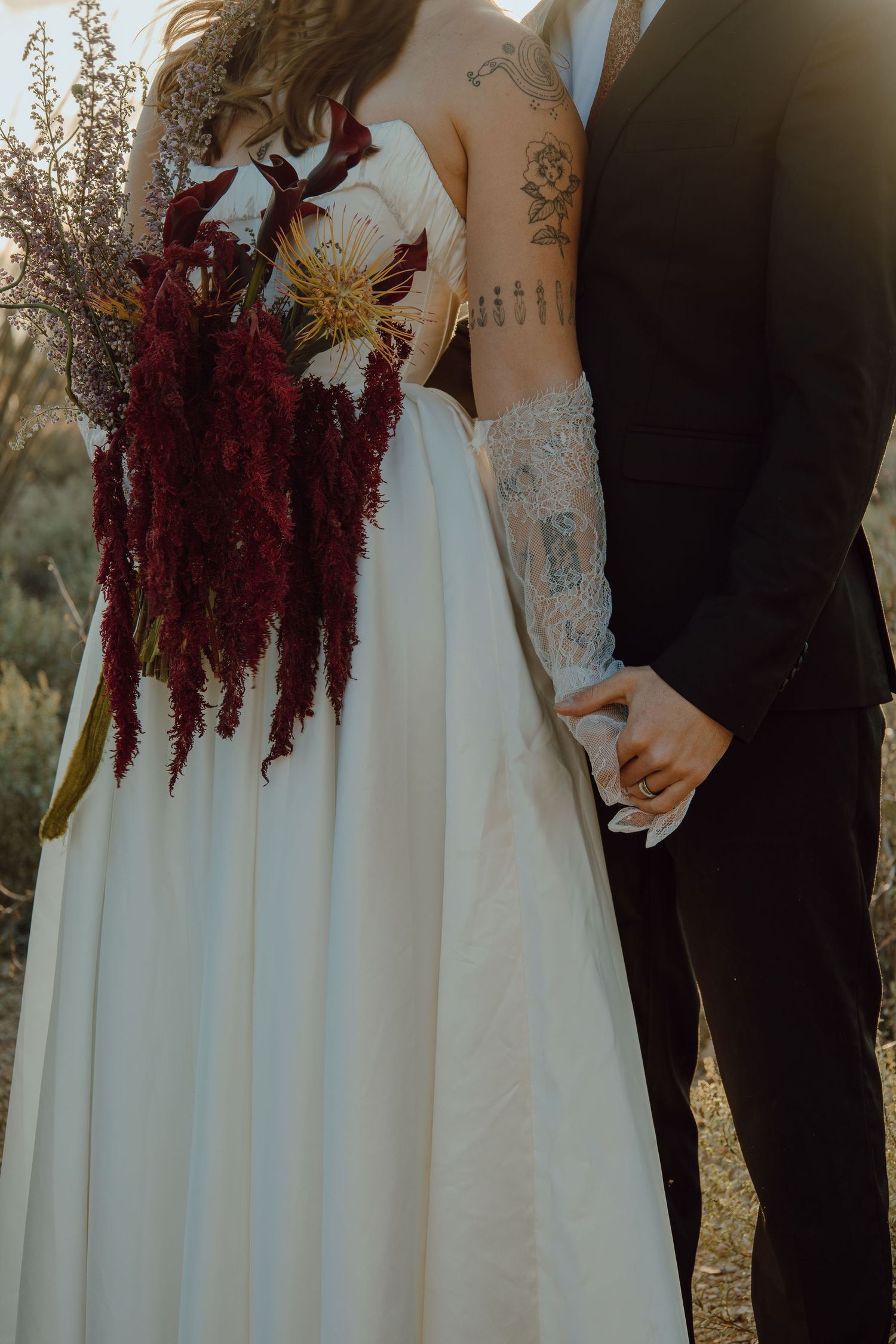 Close-up of tattooed bride and groom holding hands, bride carrying cascading burgundy floral bouquet.
