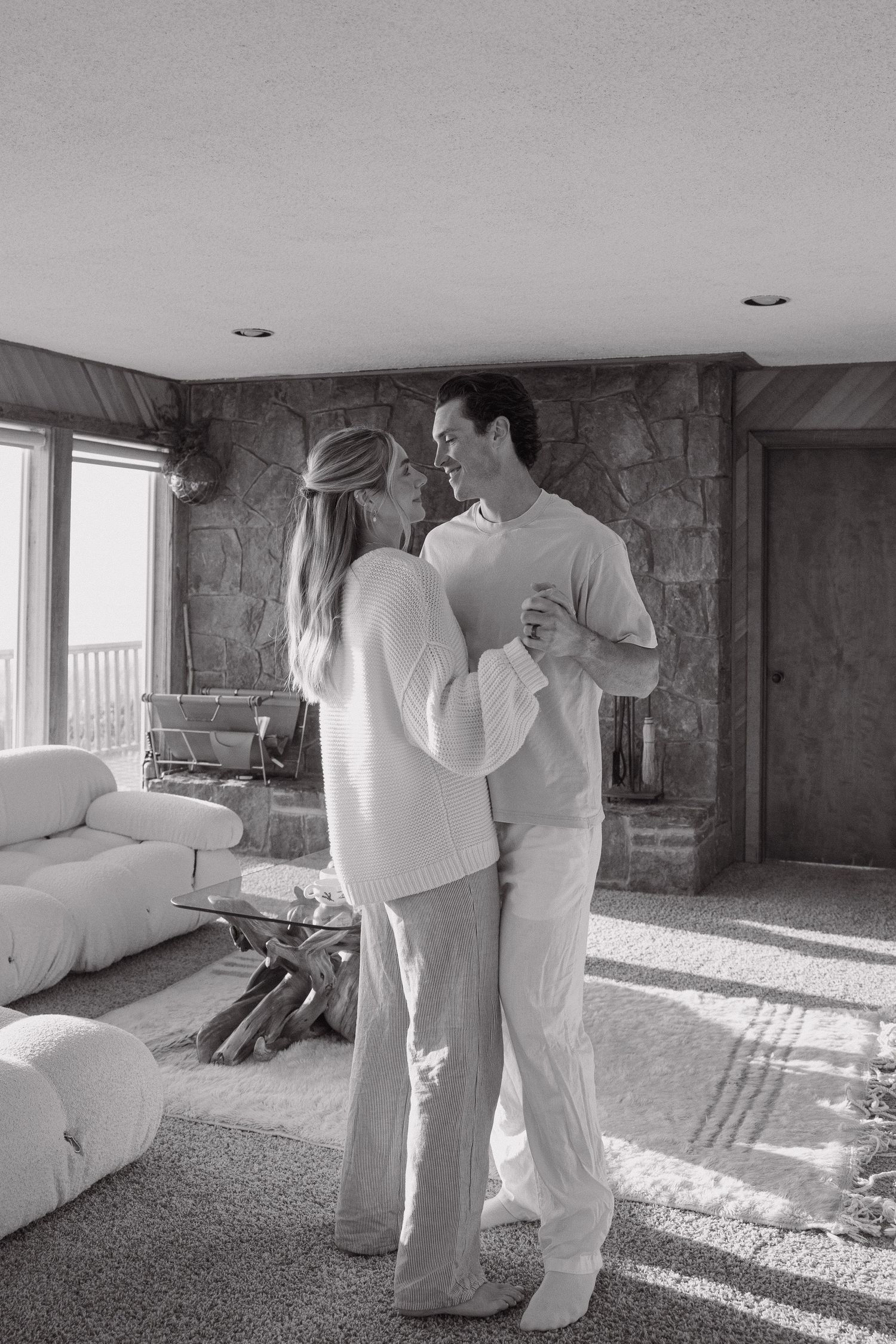 Black and white photo of a couple dancing together in a cozy living room with a stone fireplace.