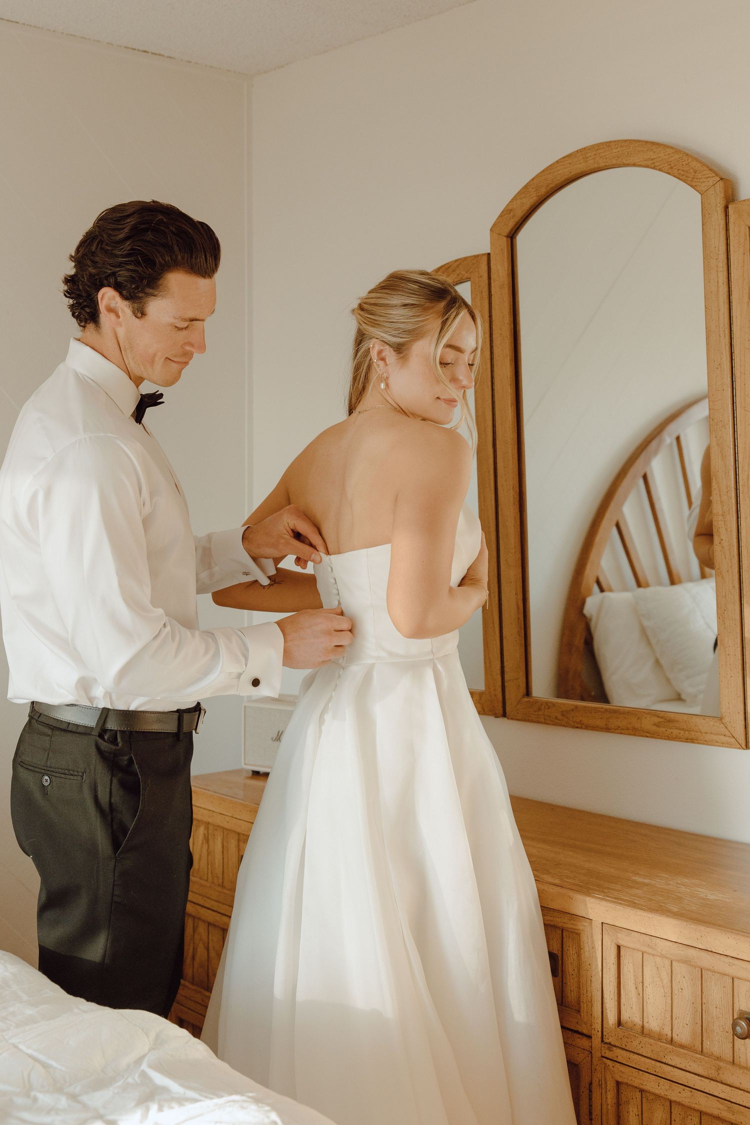 Groom in white shirt fastening the back of brides strapless white wedding gown near a wooden mirror.