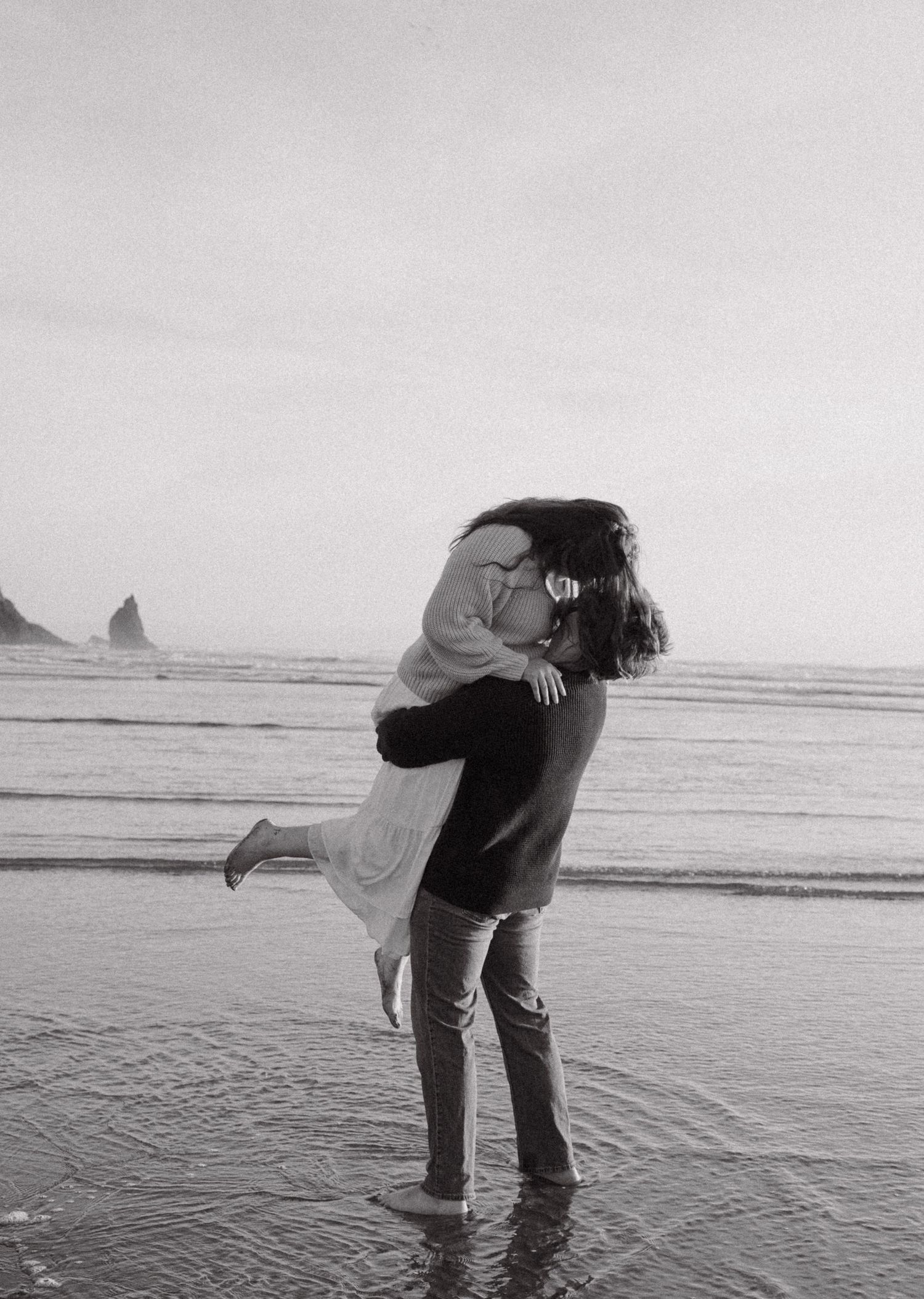 Black and white photo of a couple embracing on a beach, man lifting woman as waves wash ashore.