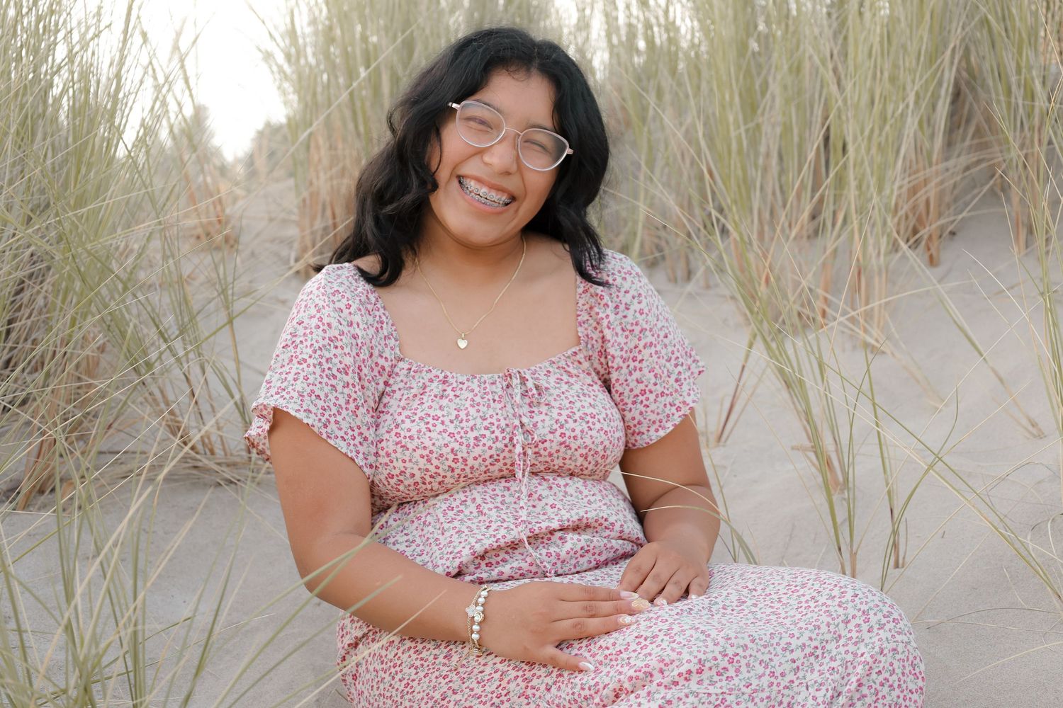 Smiling pregnant woman in floral dress sitting among beach dunes at sunset.