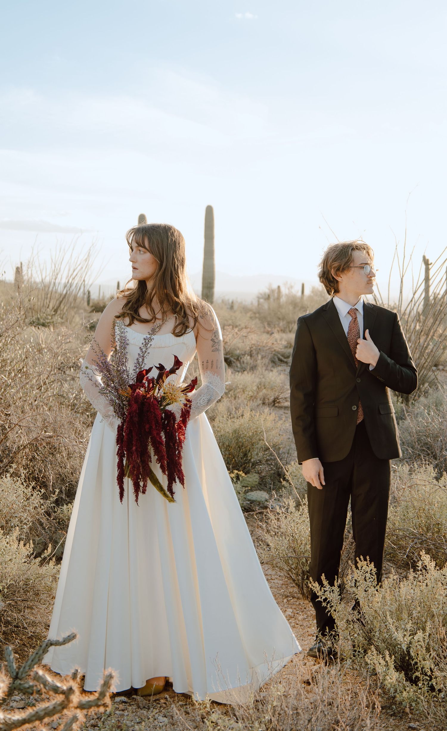 Bride in white gown and groom in dark suit pose in Sonoran desert with cacti.