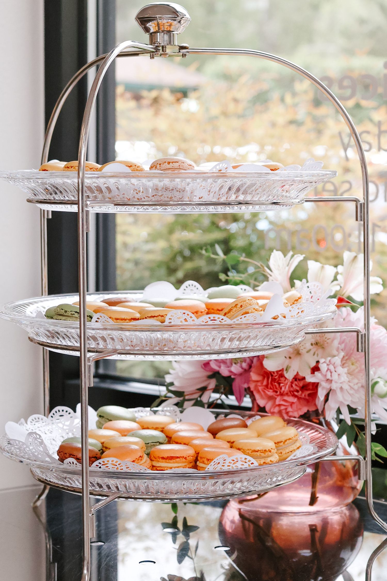 Elegant three-tier silver stand displaying colorful French macarons near flowers.