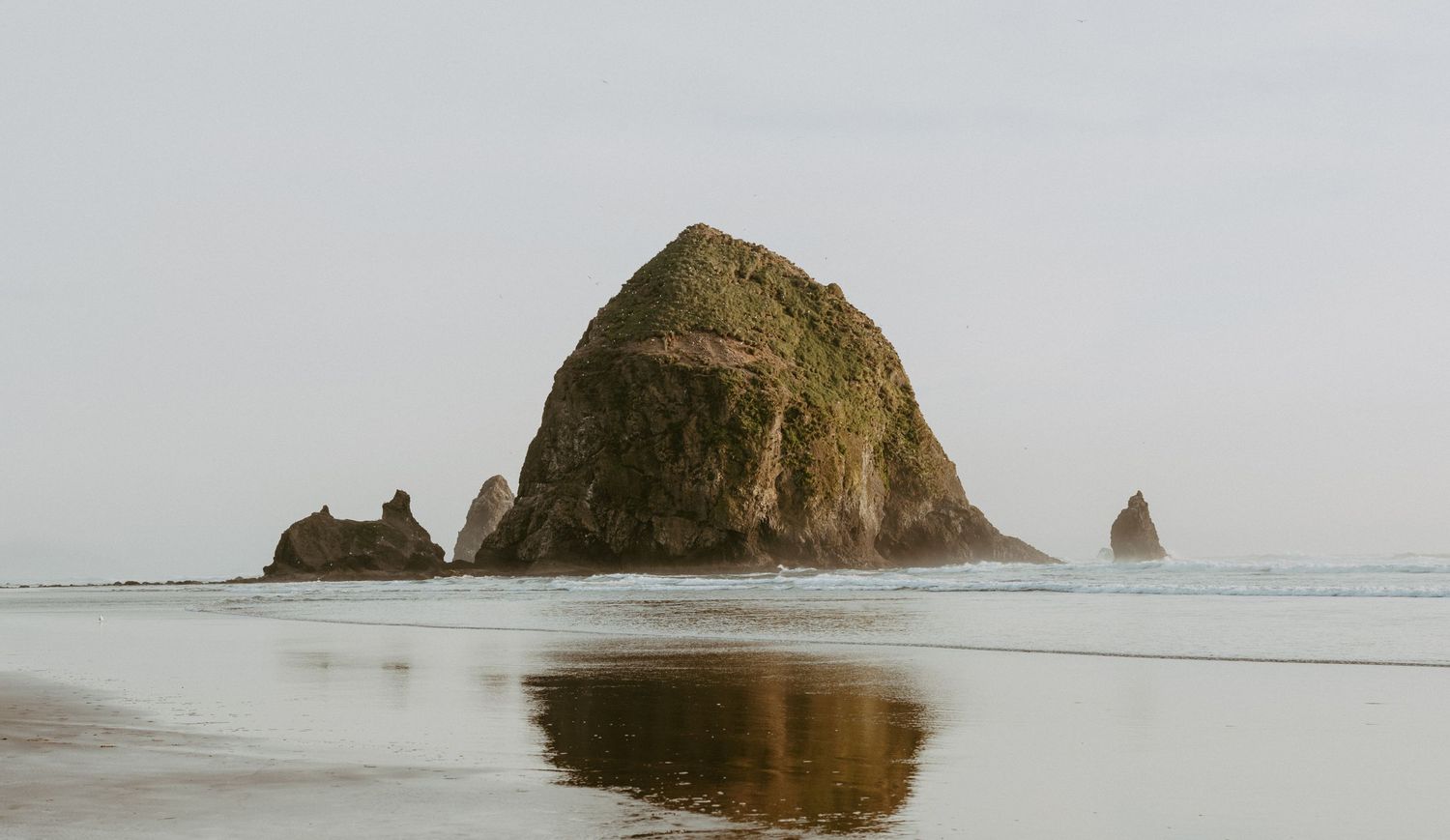 Haystack Rock reflected in wet sand at Cannon Beach, Oregon, with sea stacks visible in misty coastal waters.