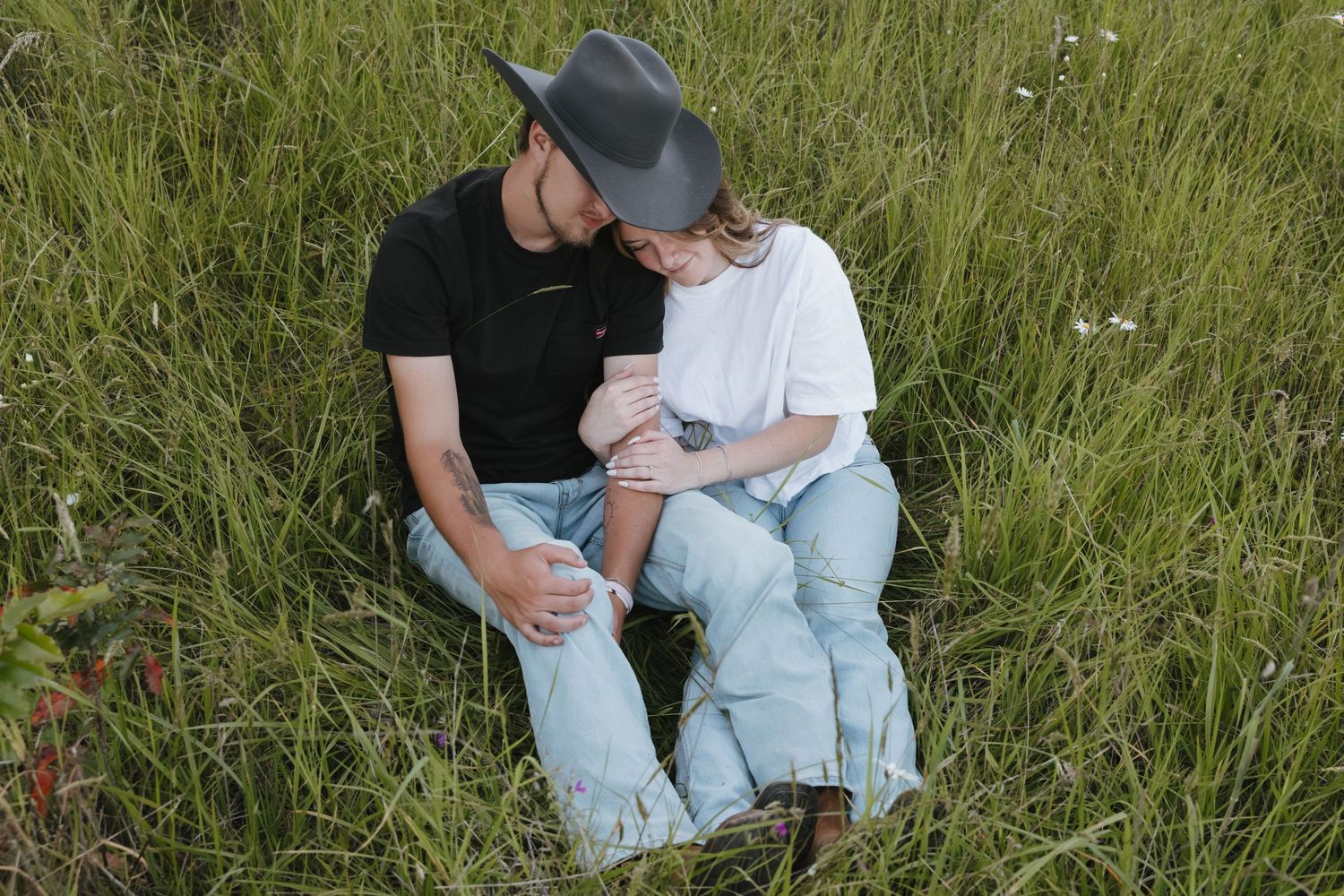 Couple sitting in green grass field, man in black tee and cowboy hat with woman in white tee and jeans.
