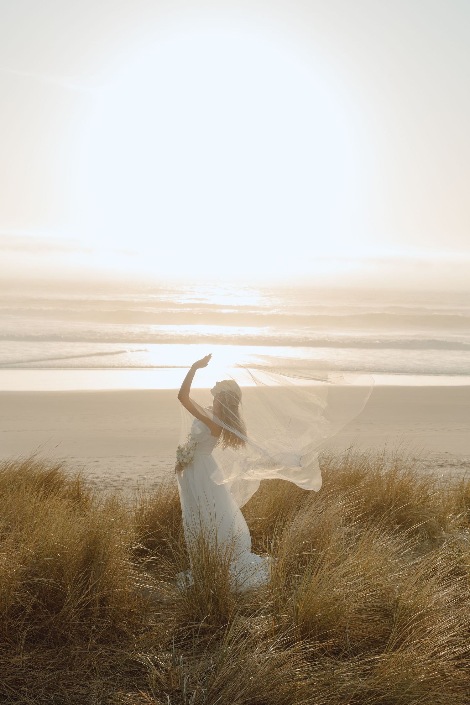 Bride in white gown and veil standing on sand dunes at sunset beach.