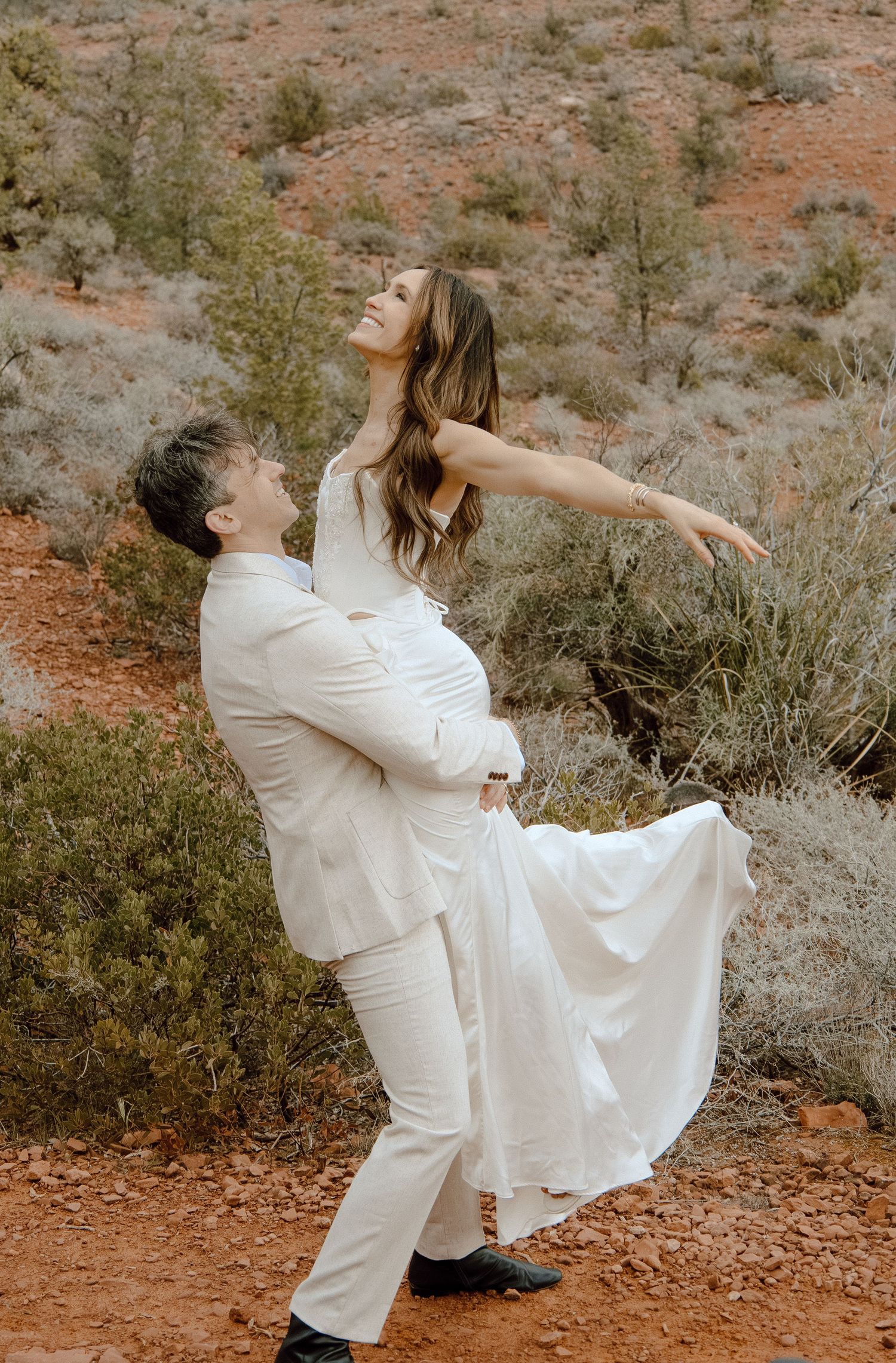 Groom lifts joyful bride in white dress on red desert trail surrounded by shrubs.