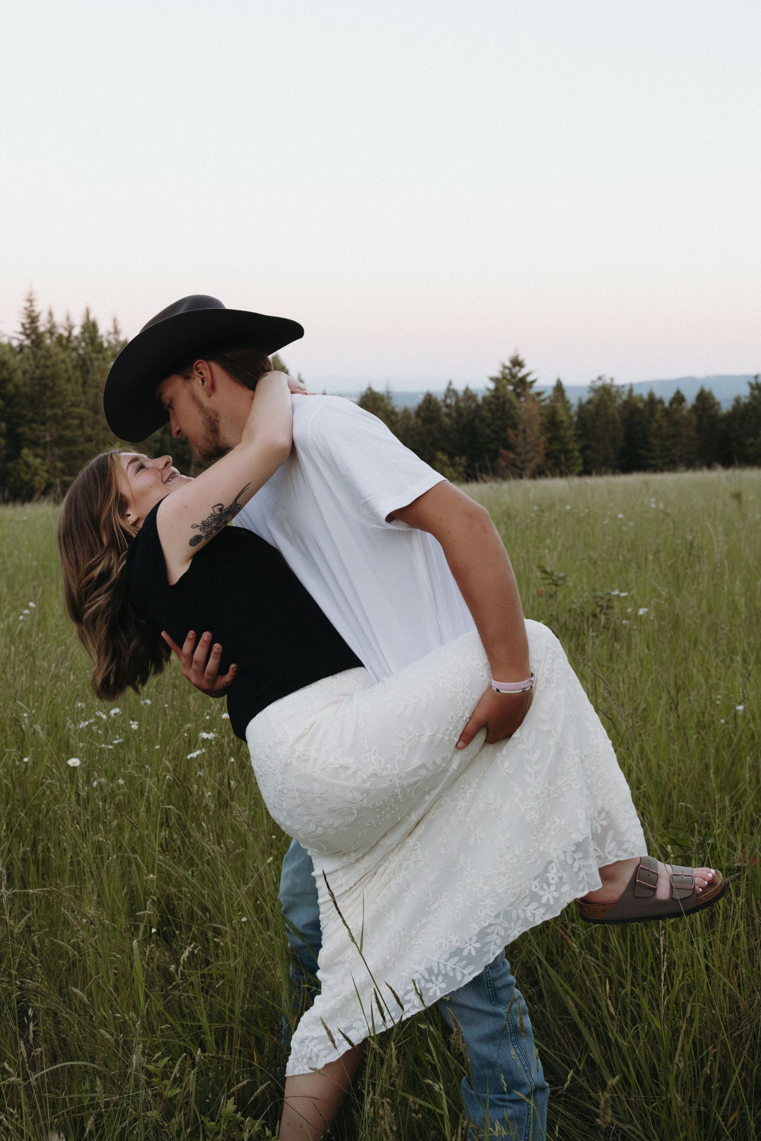 A couple sharing a romantic kiss in a meadow, man in cowboy hat lifting woman in white lace skirt.