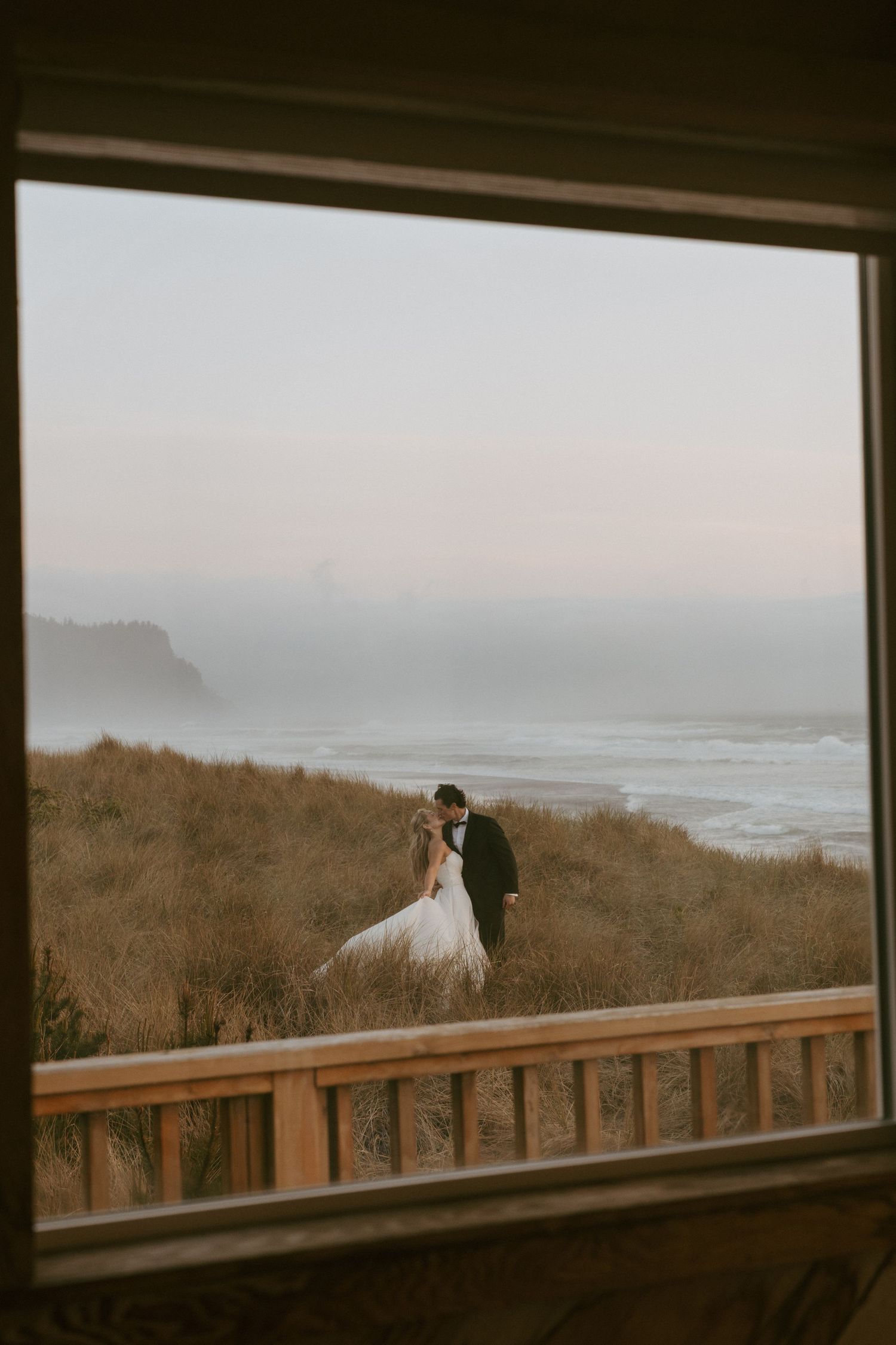 Bride and groom kissing in coastal dunes at dusk, viewed through a wooden window frame with ocean waves behind them.
