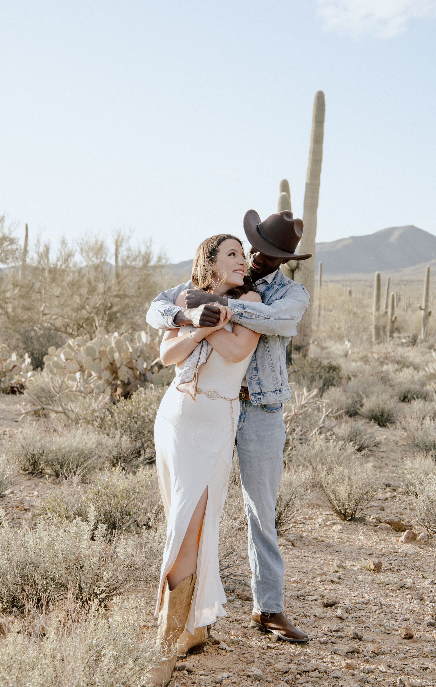 Couple embracing in Arizona desert with saguaro cacti, woman in white dress and man in cowboy hat and denim jacket.