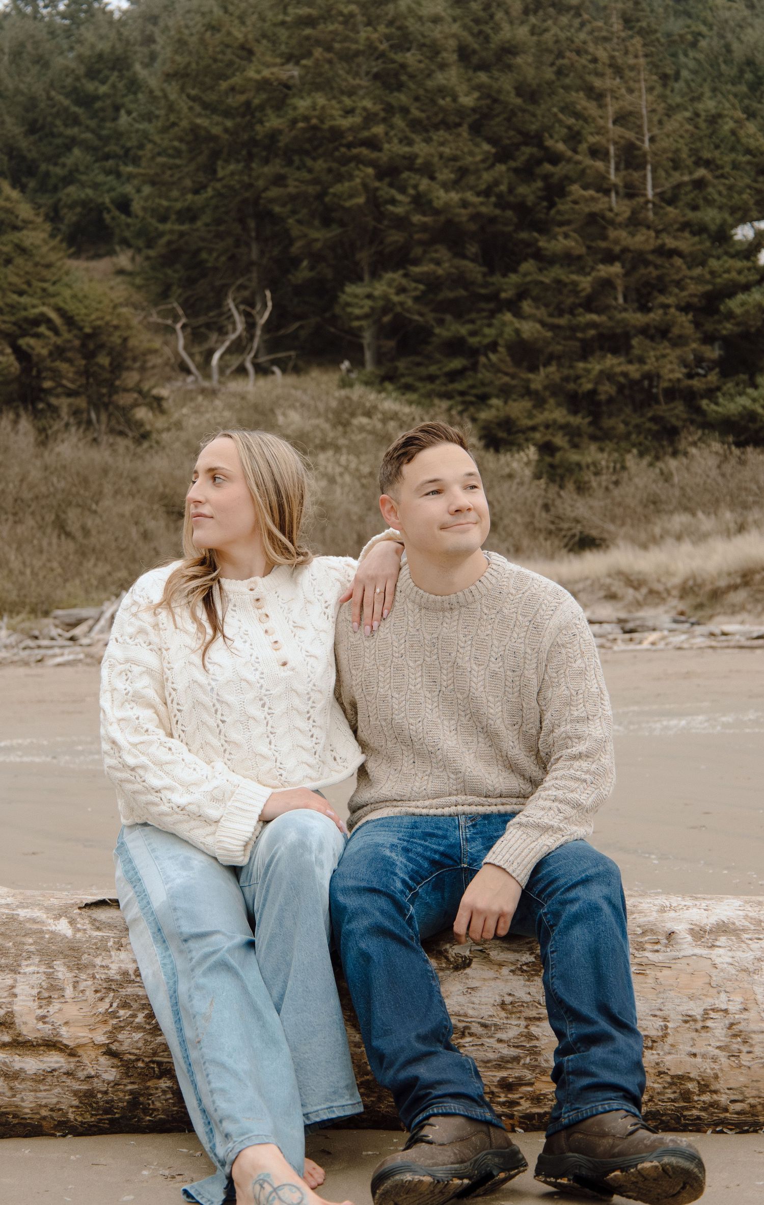 Young couple sitting on a driftwood log on a beach with lush green forest trees in the background.