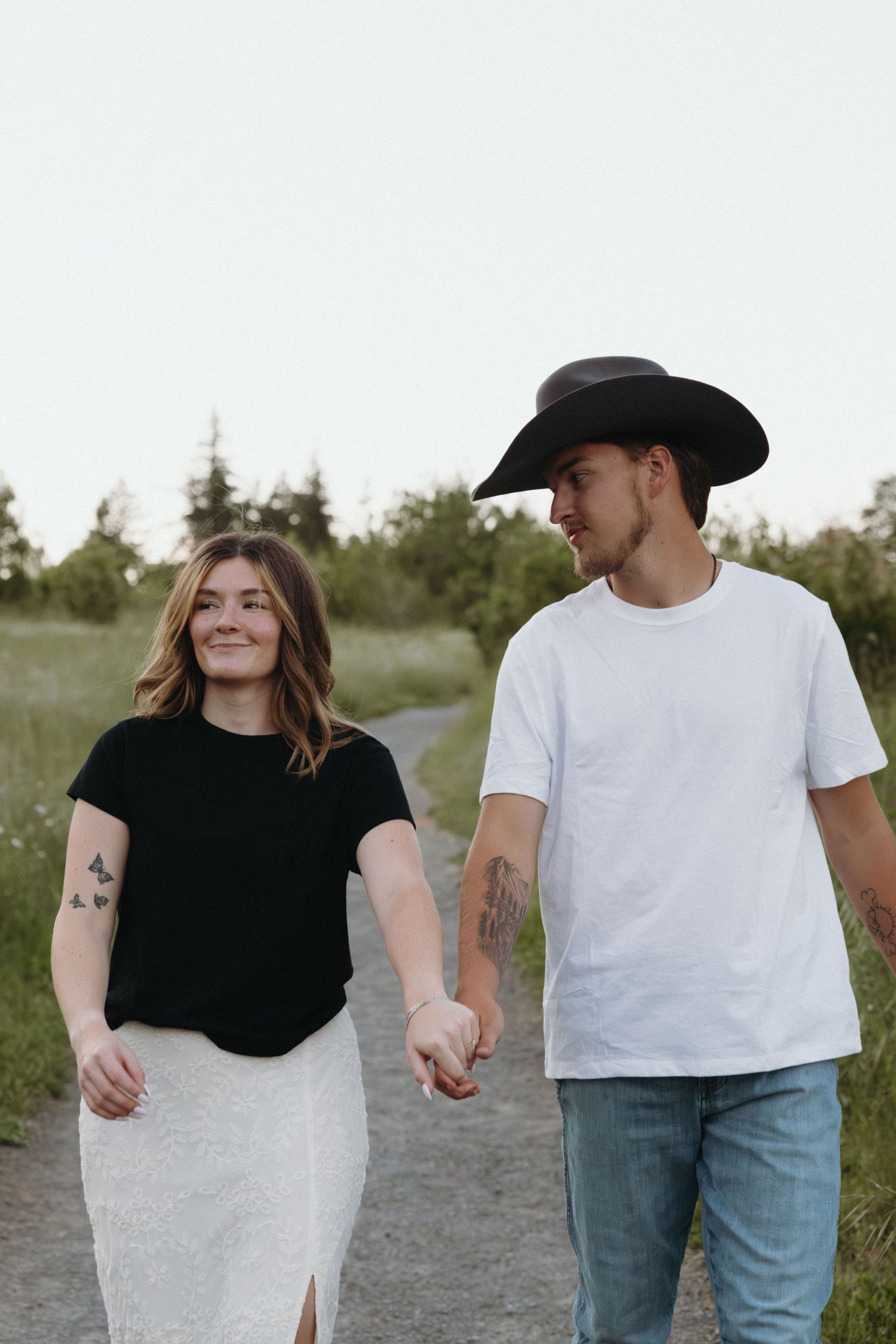 A couple holding hands and walking on a path, woman in black top and man in white tee and cowboy hat.
