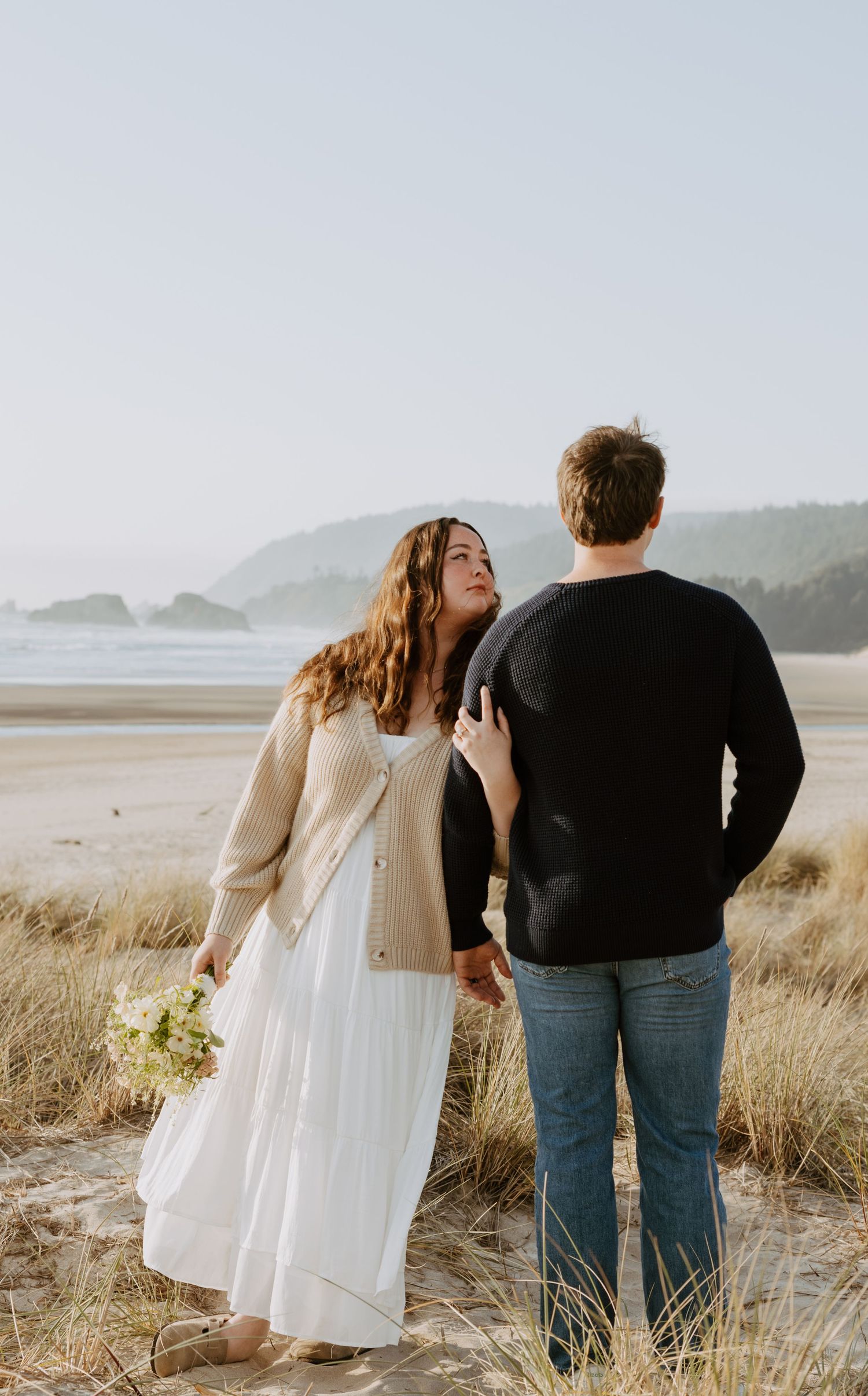 Couple holding hands on a sandy beach, woman in white dress with bouquet, man in black sweater and jeans.