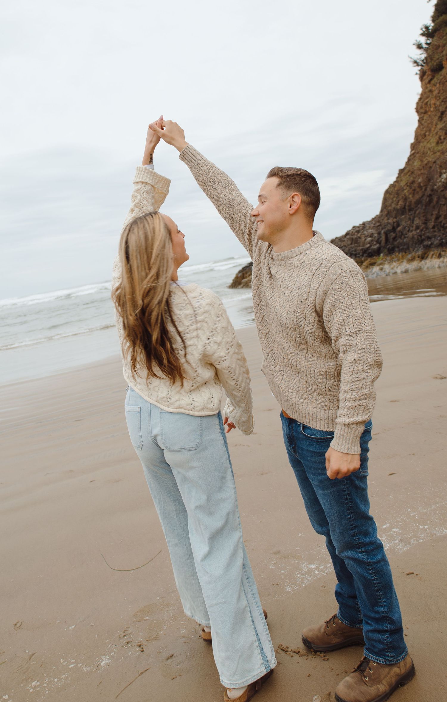 Couple dancing on a sandy beach as man twirls woman with rocky ocean cliffs in the background.