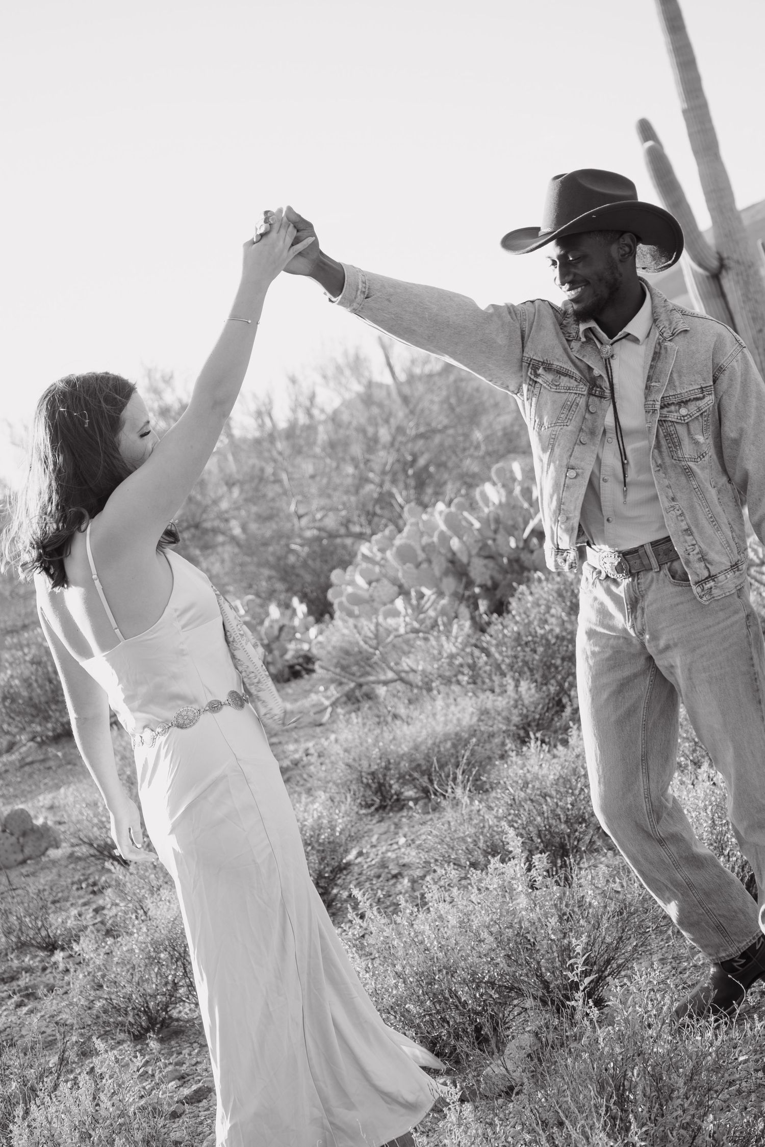 Black and white photo of couple dancing in desert landscape with saguaro cacti, man spinning woman in white dress.
