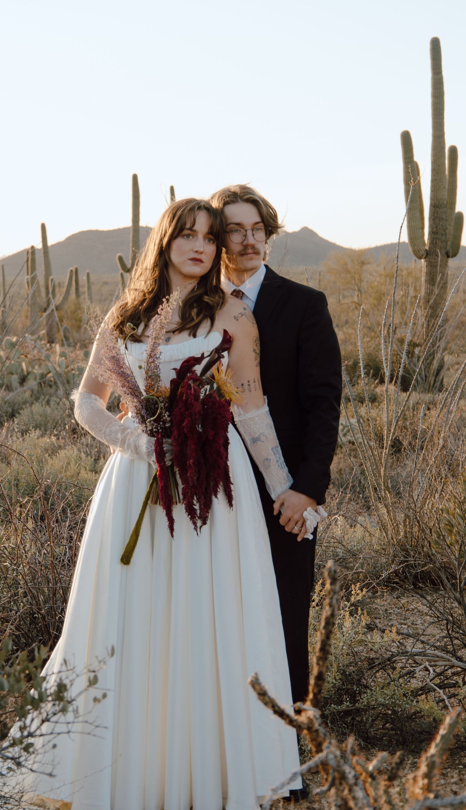 Couple in wedding attire poses among saguaro cacti in Arizona desert, bride holding deep red bouquet.