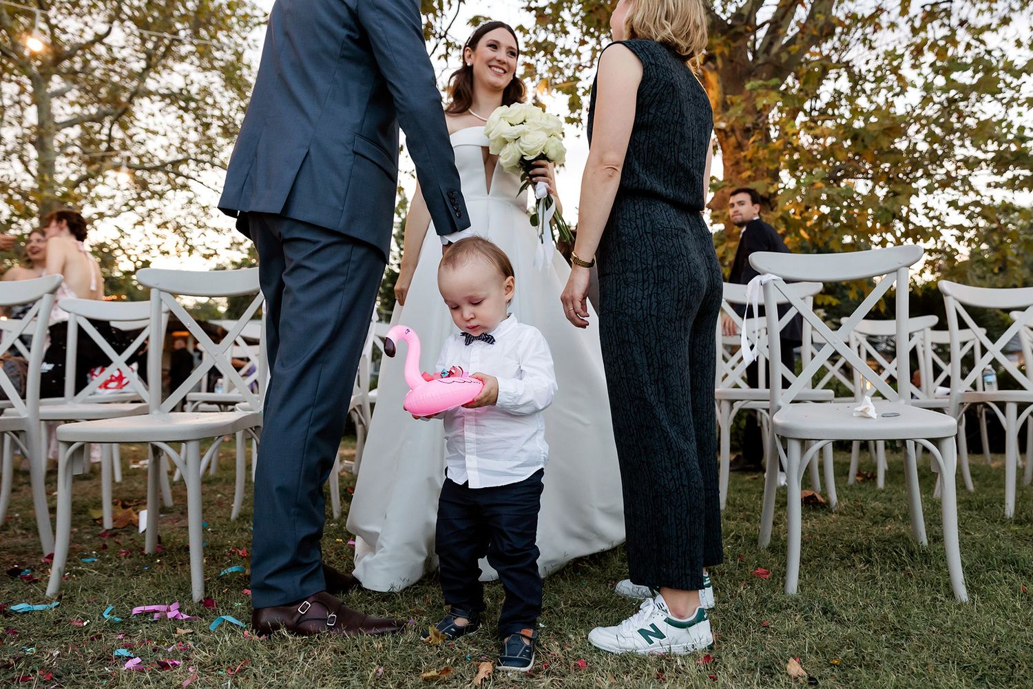 Candid documentary shot of a serious toddler holding a pink inflatable flamingo among standing adult guests at an outdoor Tekirdağ wedding.