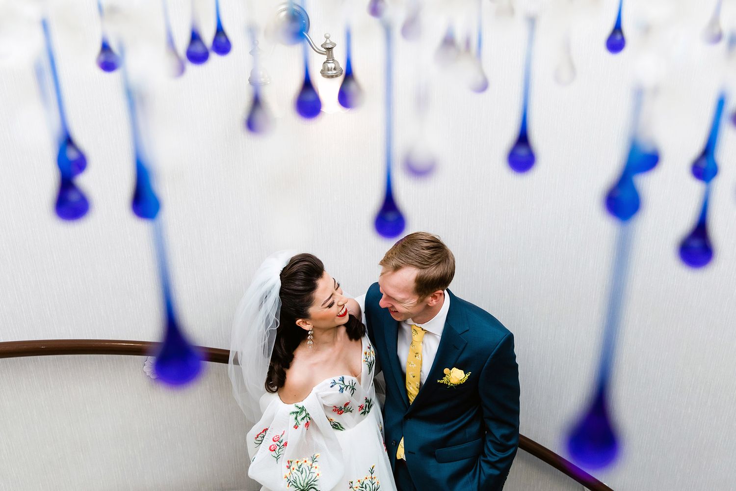 High-angle shot of a bride in a floral embroidered gown and groom in a teal suit on a spiral staircase with blue glass drops.