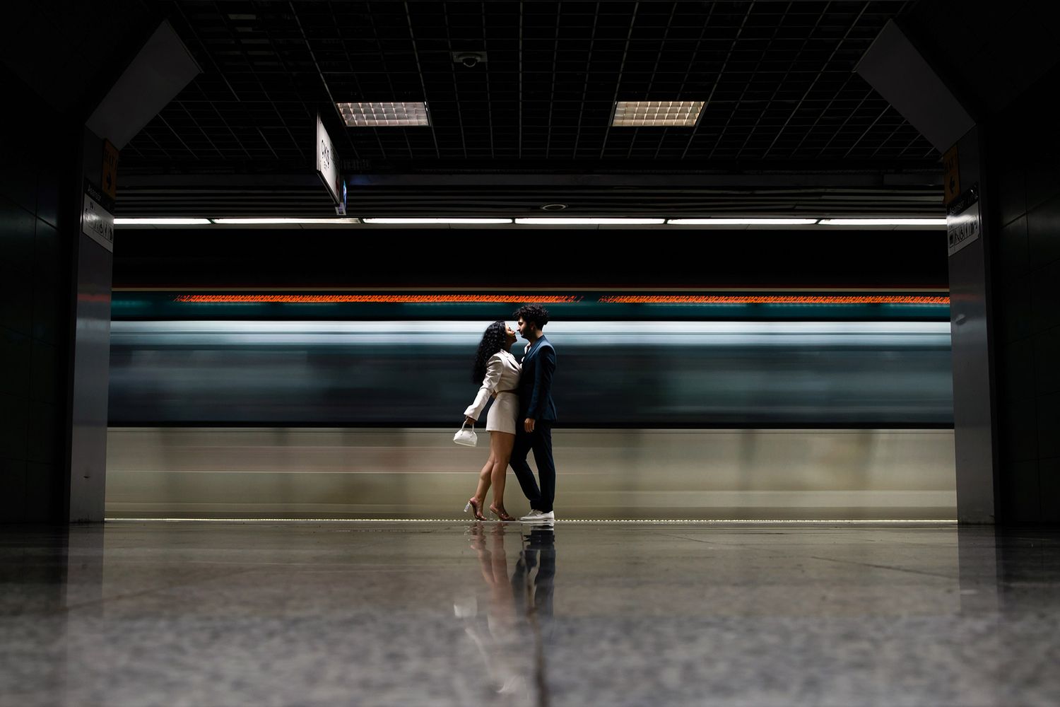Dramatic cinematic portrait of a couple standing still and embracing in Sirkeci station while a fast-moving metro train blurs behind them.
