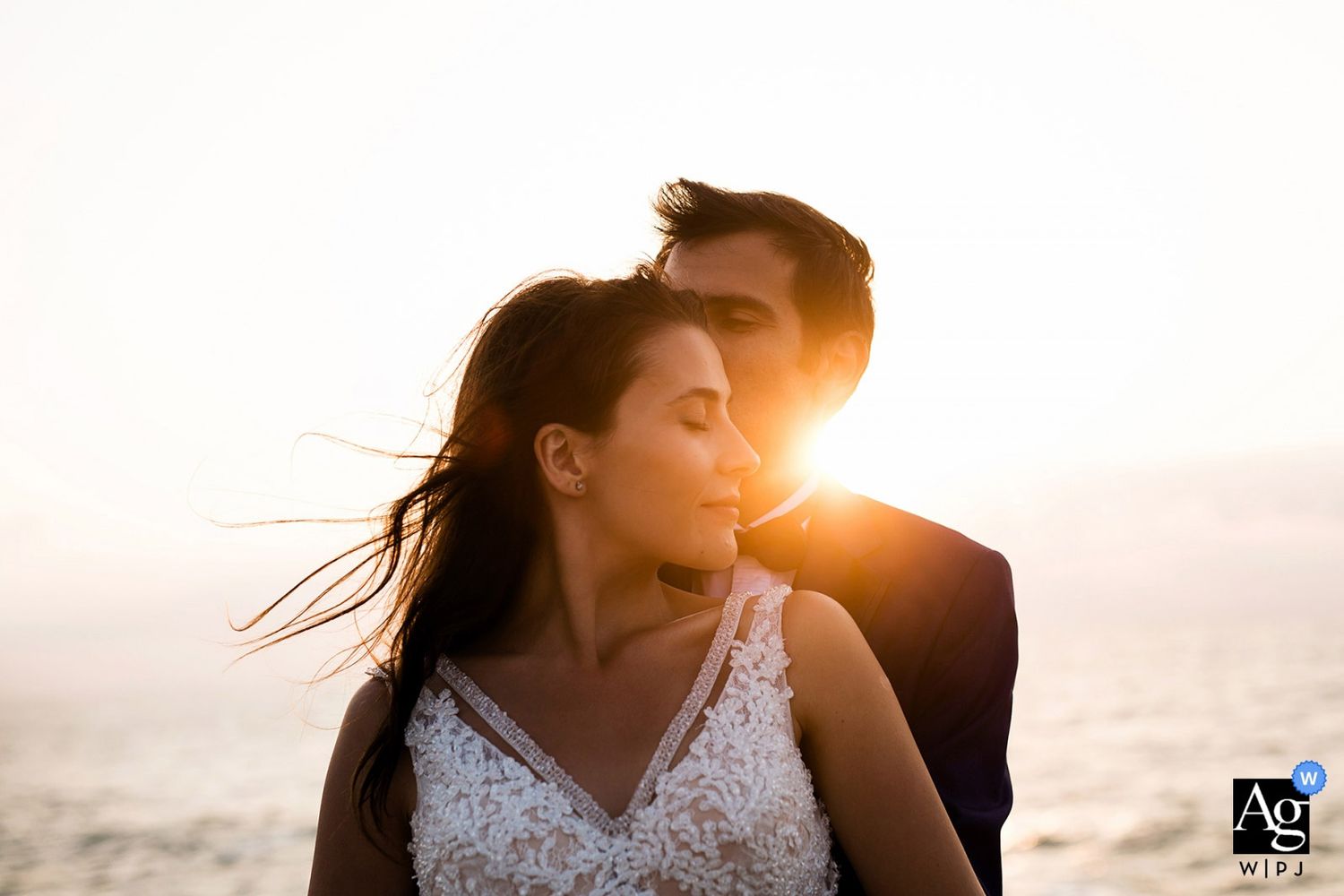 AG WPJA award-winning romantic golden hour portrait of a groom kissing his bride's cheek with beautiful sun flare and wind.