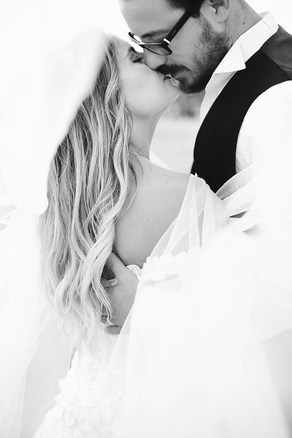 Intimate black and white documentary portrait of a bride and groom kissing, softly wrapped together underneath a flowing bridal veil.