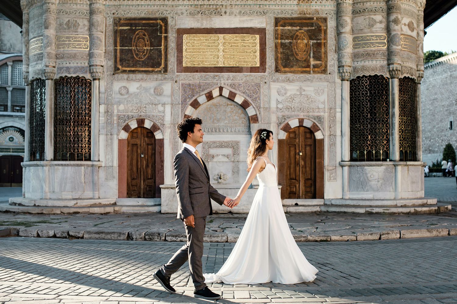 A bride and groom walk hand-in-hand past an intricate, historical marble fountain during a wedding portrait session at Topkapı Palace.