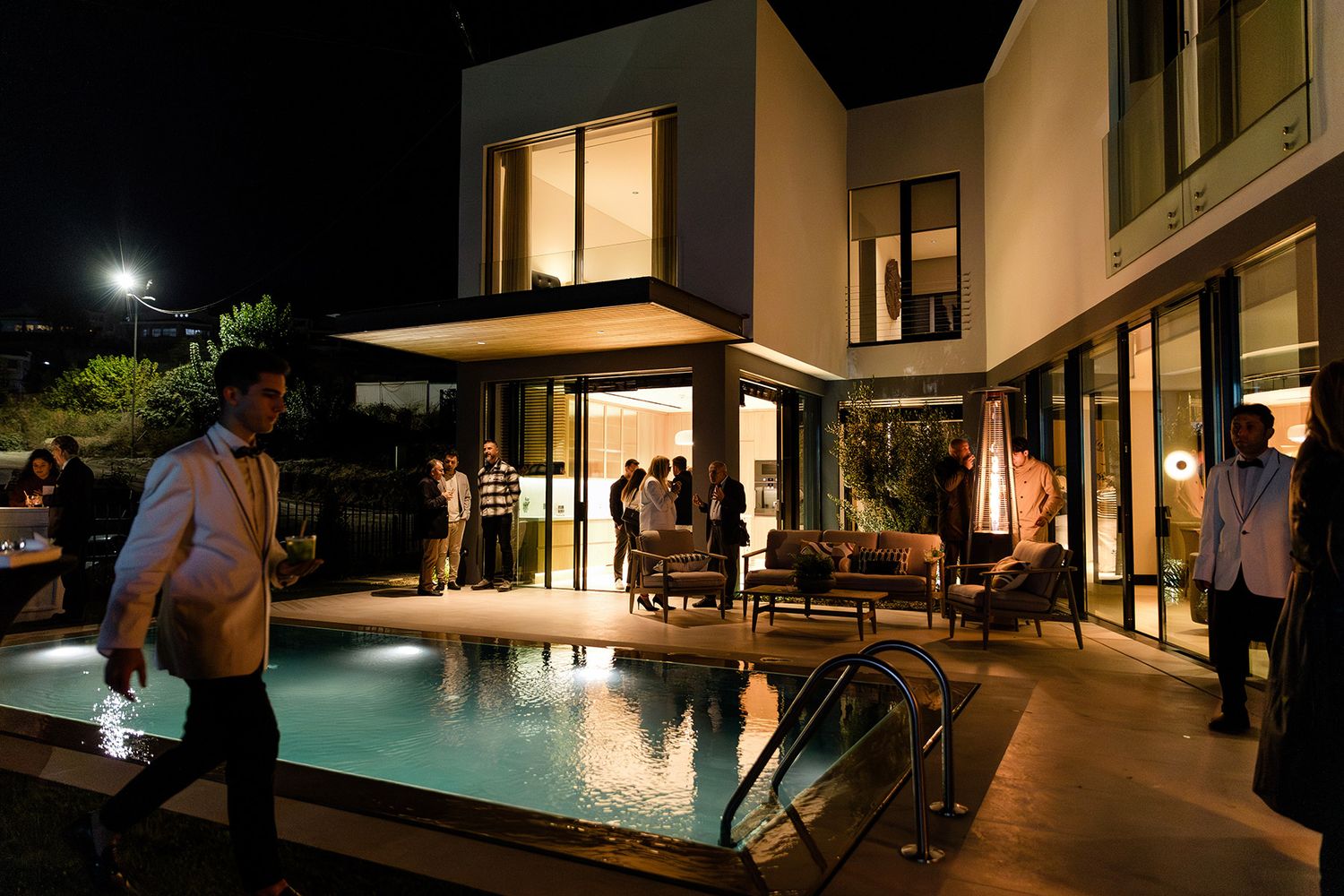 Corporate guests mingling and a waiter serving drinks at an exclusive poolside evening networking mixer at a luxury modern villa.