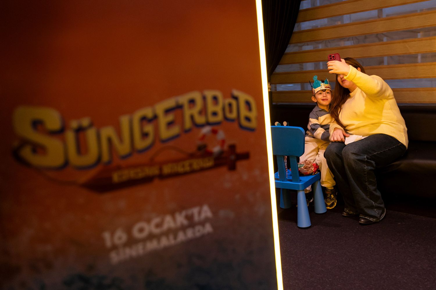 A mother taking a selfie with her child behind a brightly lit promotional sign at a SpongeBob SquarePants movie premiere brand activation.